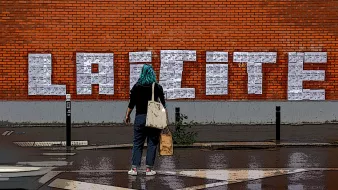 L'image montre une femme de dos, portant un foulard bleu, qui se tient devant un mur en briques rouges sur lequel est inscrit "LAICITE" en grandes lettres blanches. Le sol est humide, ce qui suggère qu'il a récemment plu. La femme porte un sac et semble contempler le graffiti sur le mur. L'environnement urbain et coloré souligne le message affiché.