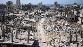 L'image montre une scène de destruction dans une zone urbaine après un conflit. De nombreux bâtiments sont partiellement détruits, avec des débris éparpillés sur le sol. On peut apercevoir des ruines de constructions, tandis qu'une route poussiéreuse traverse le paysage. À l'horizon, on voit la mer, ce qui contraste avec la gravité des destructions. L'atmosphère apparaît calme, mais la destruction est évidente et témoigne des effets dévastateurs de la guerre.