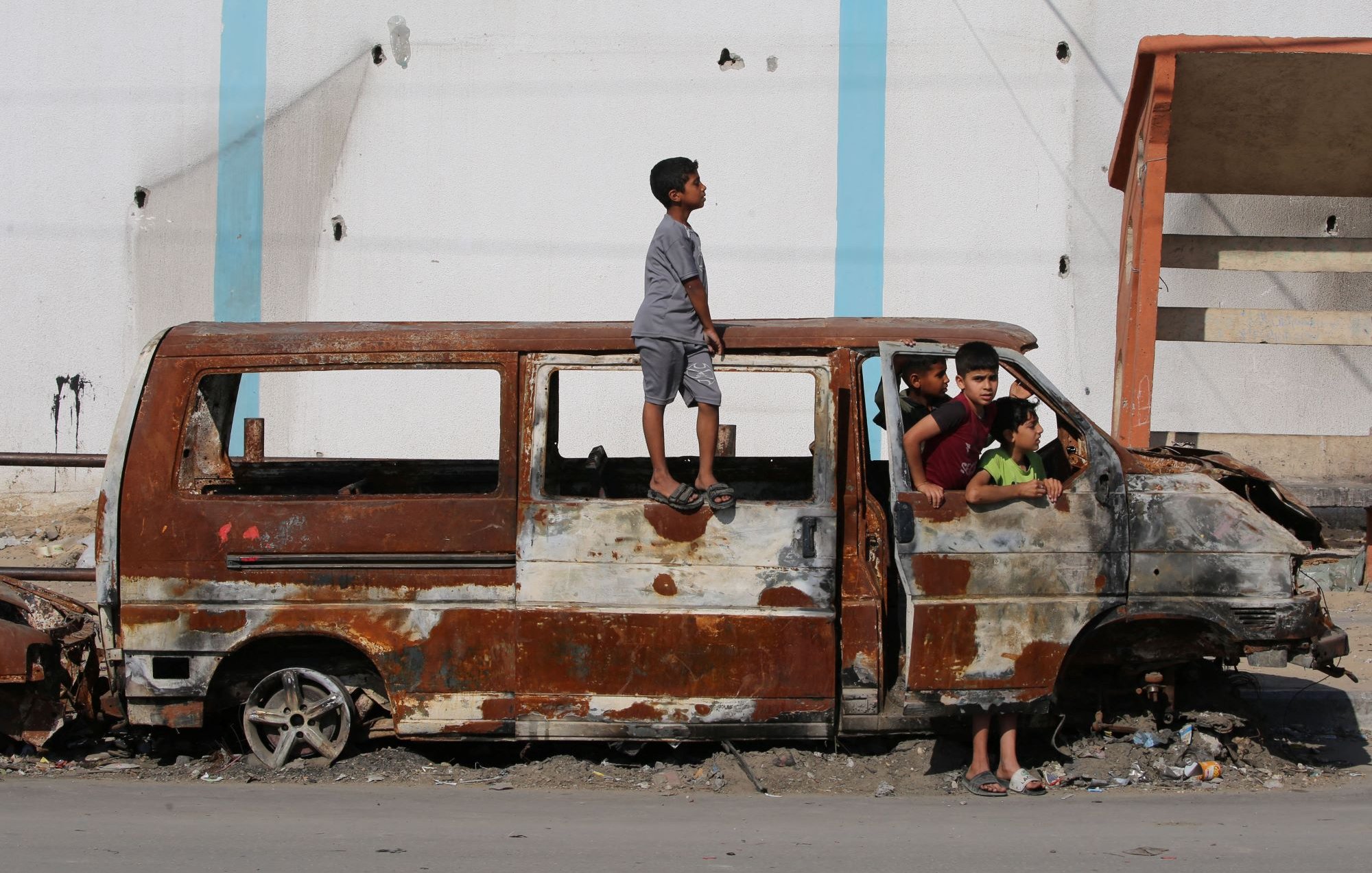 L'immagine mostra un gruppo di bambini che interagiscono attorno a un veicolo abbandonato e arrugginito. Uno dei bambini è in piedi sulla parte superiore di un furgone, mentre altri si trovano all'interno e intorno al veicolo. Lo sfondo evidenzia un edificio deteriorato, suggerendo un ambiente urbano trascurato. L'atmosfera sembra essere quella di un'area in difficoltà, ma i bambini sembrano divertirsi nonostante le condizioni.