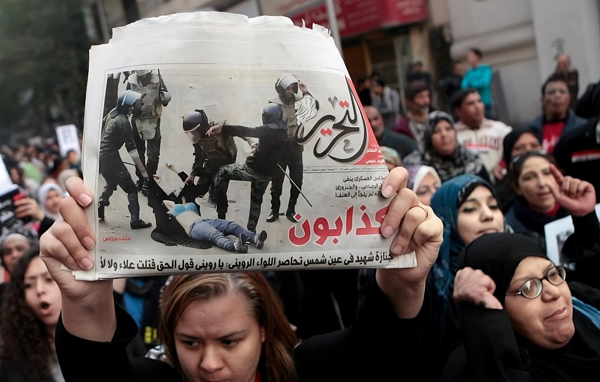 L'immagine mostra una manifestazione con un gruppo di persone unite in segno di protesta. In primo piano, una persona tiene in alto un giornale che riporta un'immagine significativa legata all'evento. La folla sembra esprimere emozioni forti, con alcune donne visibilmente agitatesi e altre che mostrano determinazione. Gli striscioni e le espressioni nei loro volti indicano il loro impegno per una causa importante. Il contesto generale evoca tensione e partecipazione attiva a una questione sociale.