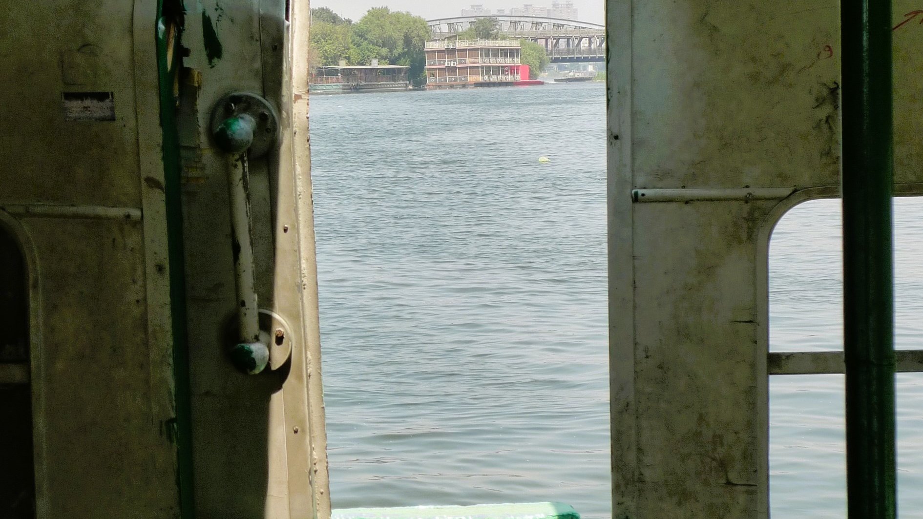 L'immagine mostra l'interno di un'imbarcazione, con una porta aperta che si affaccia su un corpo d'acqua. La vista rivela un paesaggio sereno, con un riflesso dell'acqua e vegetazione visibile in lontananza. La struttura della barca appare semplice, con pareti in metallo e sedili visibili all'interno. La luce naturale entra attraverso l'apertura, creando un'atmosfera tranquilla.
