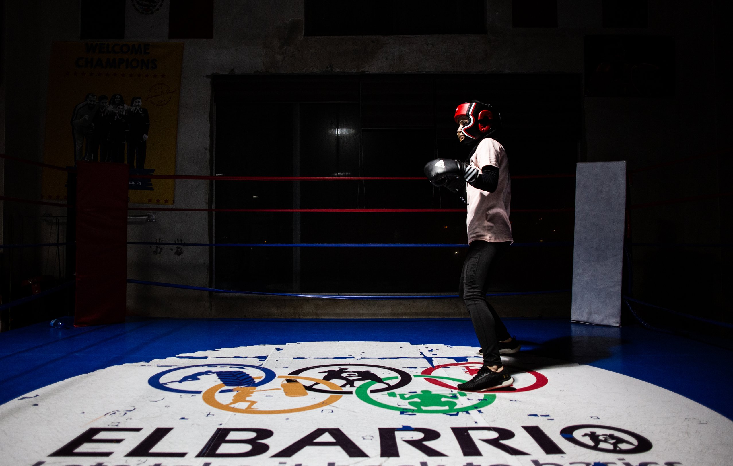 L'immagine mostra un giovane pugile che indossa i guantoni e un casco protettivo, mentre si allena in un ring da boxe. Lo sfondo è parzialmente illuminato, mettendo in risalto il pugile in azione. Sotto di lui, si trova un logo con i simboli olimpici e la scritta "ELBARRIO", che suggerisce un ambiente dedicato allo sport e alla formazione. La scena ha un'atmosfera di concentrazione e determinazione.