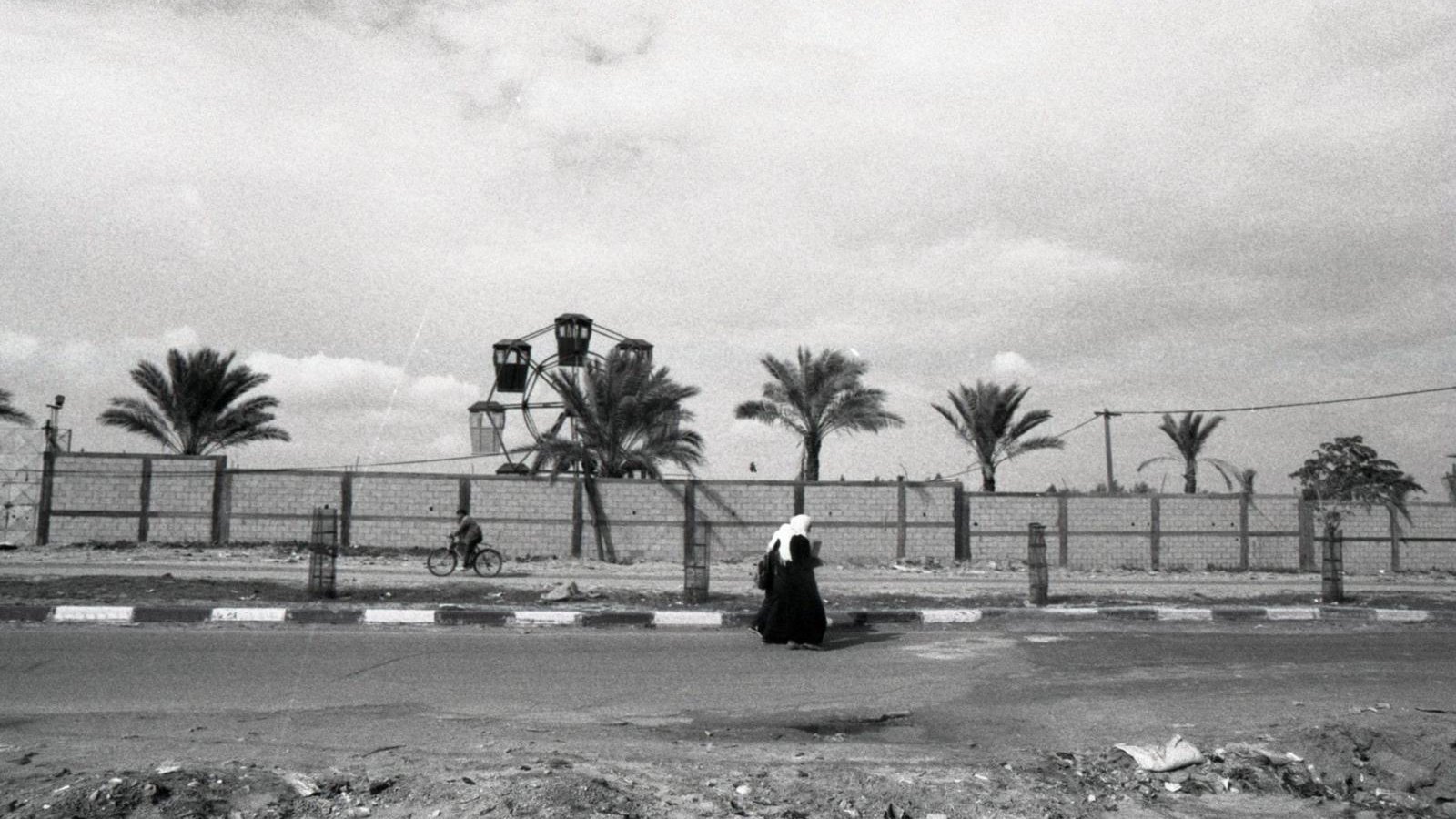 L'immagine ritrae una scena in bianco e nero che mostra una figura solitaria che cammina lungo una strada. Sullo sfondo, si possono vedere alcune palme e una ruota panoramica, che suggerisce la presenza di un parco o di un'area ricreativa. La composizione offre un'atmosfera malinconica e riflessiva, accentuata dalla scelta dei toni scuri e dalla mancanza di colori vivaci. La figura, vestita con abiti tradizionali, sembra essere in un momento di solitudine all'interno di un paesaggio urbano.