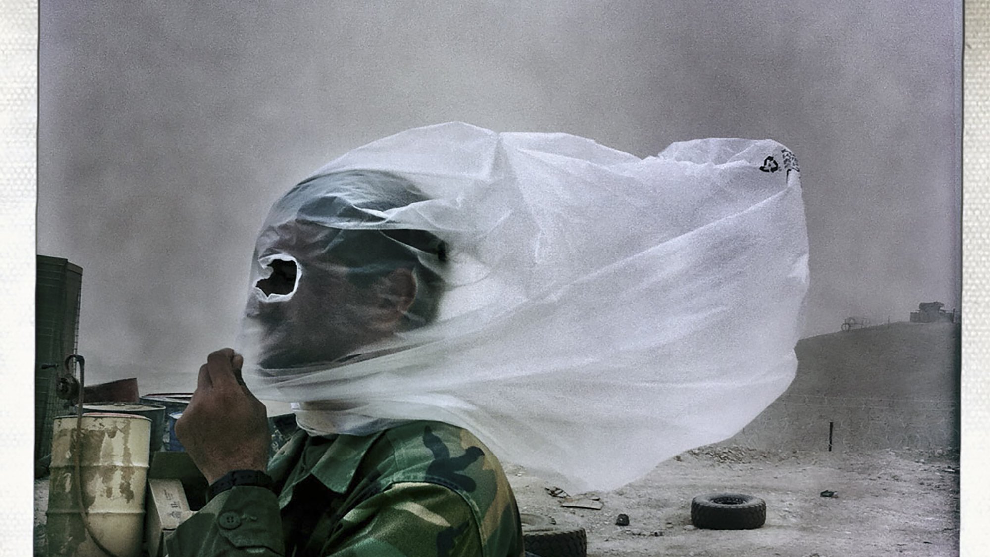 L'immagine mostra un uomo in uniforme militare, avvolto da un velo di stoffa che sembra fluttuare nel vento. Il suo viso è parzialmente coperto, rendendolo quasi irriconoscibile. Sullo sfondo si intravede un paesaggio desolato, con sabbia e detriti, suggerendo che si trovi in un ambiente arido, probabilmente in una zona di conflitto. L'atmosfera appare drammatica a causa del vento che solleva la polvere.