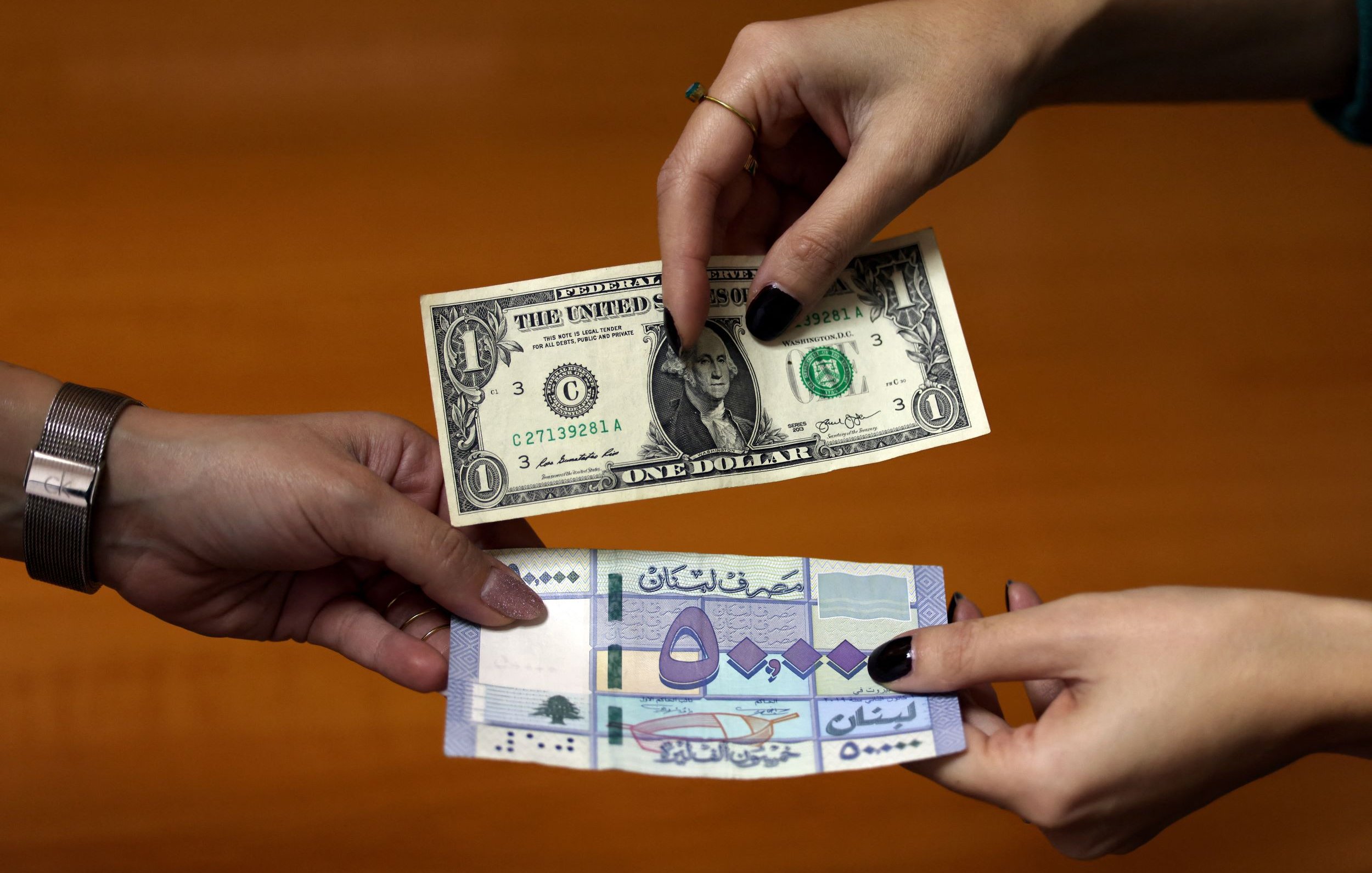 L'immagine mostra due mani che si scambiano delle banconote. Una mano tiene una banconota da un dollaro statunitense, mentre l'altra mano presenta una banconota di una valuta diversa, probabilmente una banconota di un paese arabo. Il fondo sembra essere un tavolo di legno, creando un contrasto con le banconote in primo piano.