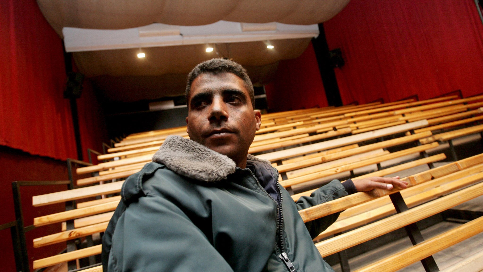 L'immagine mostra un giovane uomo seduto su una panca di legno in un teatro. Il teatro ha un'illuminazione soffusa, con tende rosse che coprono le pareti e un palcoscenico visibile sullo sfondo. L'uomo indossa una giacca scura con un colletto di pelliccia. La sua espressione sembra seria e contemplativa mentre guarda verso l'esterno, evidenziando un'atmosfera di introspezione nel contesto di un luogo culturale. Le file di sedili vuoti suggeriscono un ambiente che sta aspettando un evento o una performance.