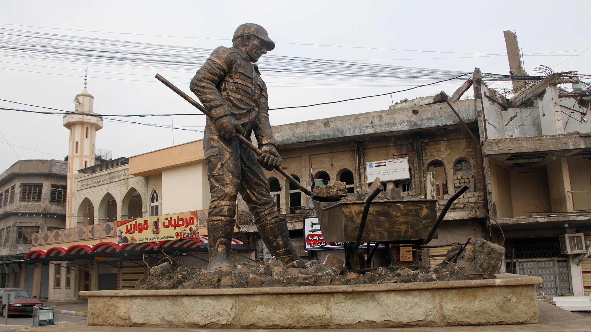 L'immagine mostra una statua di un operaio che lavora, con una pala in mano e una carriola accanto a lui. Il monumento è situato in una zona urbana, caratterizzata da edifici danneggiati e in rovina sullo sfondo. La scena trasmette un senso di resilienza e dignità, riflettendo la vita e il lavoro in un contesto difficile. L'atmosfera è un po' desolata a causa delle strutture danneggiate, mentre la statua rappresenta il duro lavoro e l'impegno quotidiano delle persone.