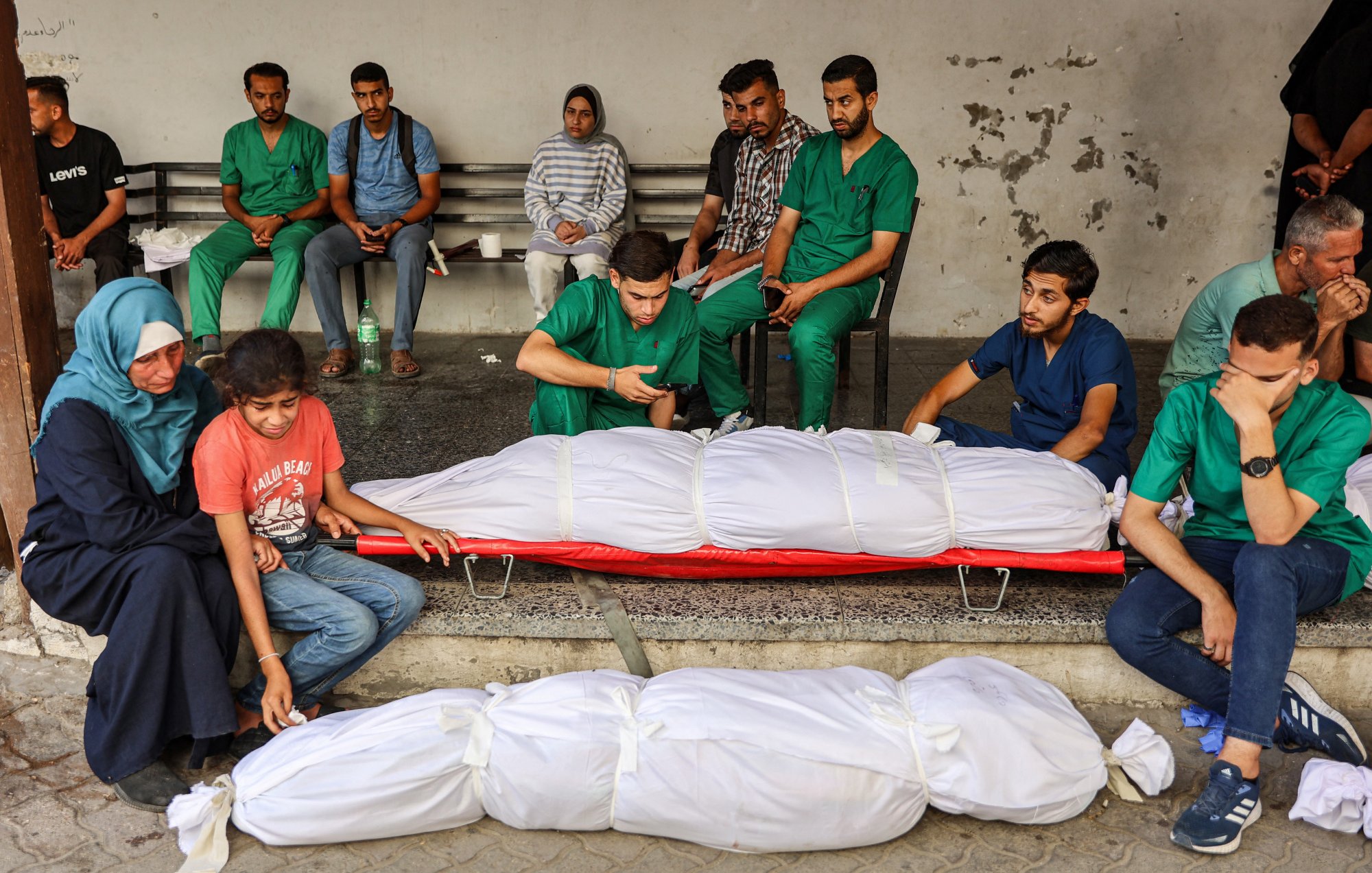 L'immagine mostra un gruppo di persone in uno spazio pubblico. Alcuni individui indossano camici verdi, suggerendo che potrebbero essere operatori sanitari. In primo piano, ci sono due corpi avvolti in lenzuola bianche. Alcuni membri del gruppo sembrano essere in uno stato di lutto, mentre altri osservano la scena in modo serio. L'atmosfera complessiva è di tristezza e riflessione.