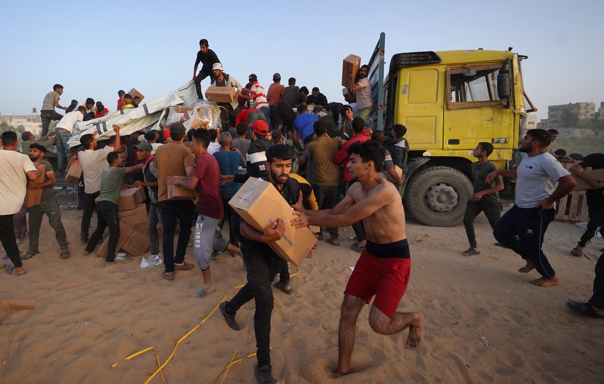 L'immagine mostra un gran numero di persone, principalmente giovani uomini, che sono radunati attorno a un camion giallo. Alcuni di loro sembrano scaricare scatole, mentre altri stanno trasportando i pacchi. L'ambiente appare polveroso e le persone sono visibilmente attive e impegnate nell'operazione di distribuzione, con espressioni di urgenza. Alcuni individui sono a torso nudo e indossano abiti vari. Si percepisce un'atmosfera di collaborazione nel tentativo di ricevere e distribuire i rifornimenti.