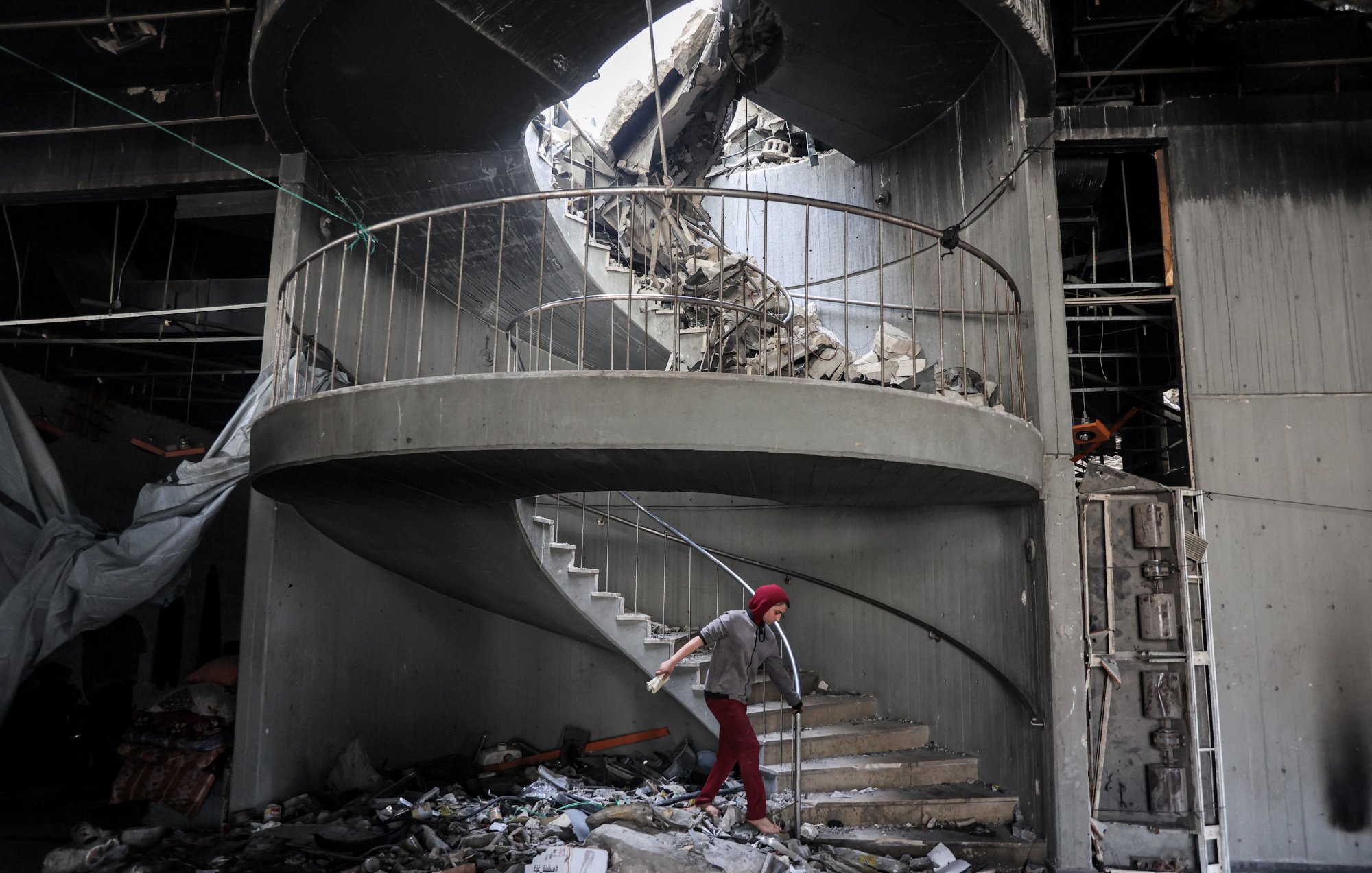 L'immagine mostra una scala a chiocciola danneggiata all'interno di un edificio in rovina. La struttura presenta macerie sparse e parte del soffitto è crollato, creando un'apertura sopra la scala. Una persona, vestita con un abbigliamento rosso e coperta da un copricapo, sta camminando sulla scala tra le macerie. L'atmosfera è quella di un luogo in crisi, con segni evidenti di distruzione e abbandono.