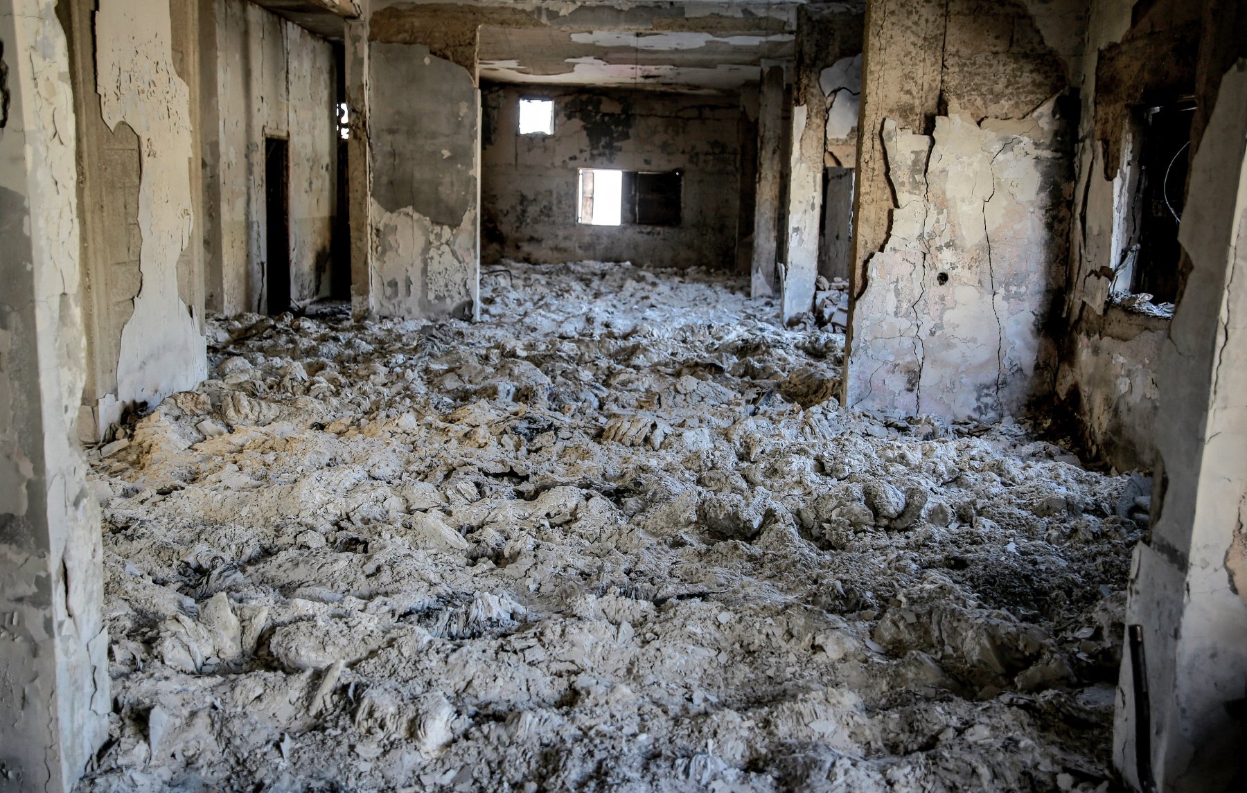 L'immagine mostra un interno di un edificio gravemente danneggiato. Le pareti sono deteriorate e presentano crepe, mentre il soffitto appare scrostato. Il pavimento è coperto da una massa bianca, che potrebbe essere polvere o detriti. I fori nelle pareti e nelle finestre suggeriscono che l'area è stata abbandonata e distrutta, creando un'atmosfera di desolazione e degrado. La luce che entra attraverso le aperture crea un contrasto con le ombre all'interno dello spazio.