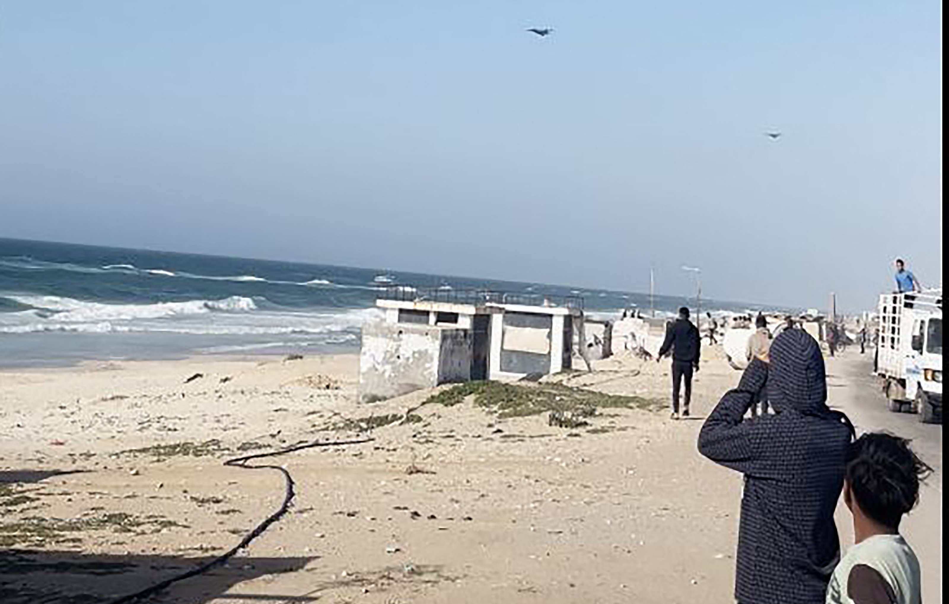 L'immagine mostra una scena costiera con il mare e delle onde in movimento. Si possono vedere alcune persone sulla spiaggia che osservano il mare, apparentemente interessate a qualcosa in cielo. Il paesaggio è caratterizzato da sabbia chiara e una scalinata che porta a una struttura, mentre in lontananza si vedono veicoli e persone che si muovono lungo la costa. Il cielo è sereno, con poche nuvole.