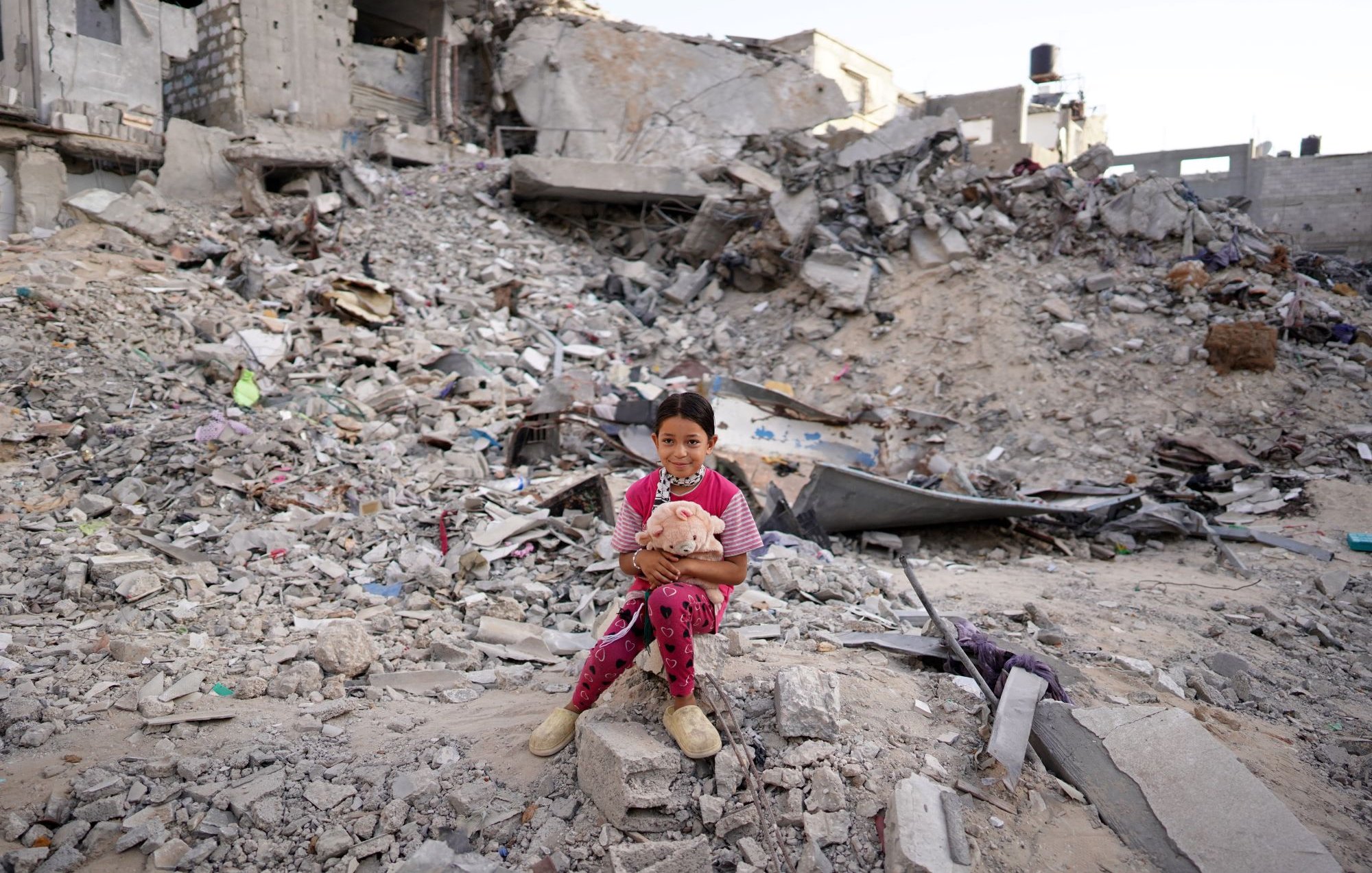 L'immagine ritrae una bambina seduta su un cumulo di macerie, circondata da un paesaggio distrutto. Il suo sguardo è diretto verso la fotocamera, mentre tiene un peluche tra le braccia. Indossa un pigiama a pois rosa e sembra essere in un ambiente devastato, con edifici in rovina sullo sfondo. La scena trasmette una forte emozione, evidenziando la vulnerabilità e la resilienza della giovane ragazza in un contesto difficile.