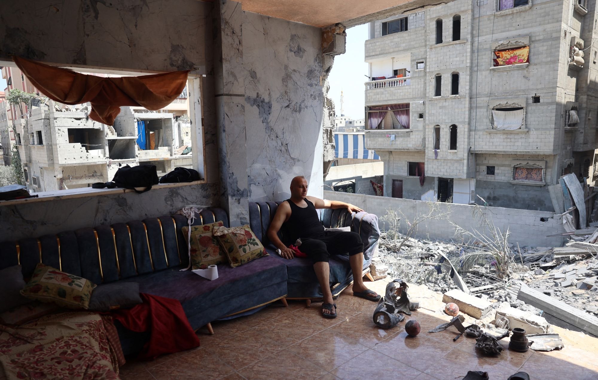 L'immagine mostra un uomo seduto su un divano in un ambiente che appare distrutto. Le pareti sono danneggiate e si possono vedere segni evidenti di distruzione. Nella stanza ci sono alcuni oggetti sparsi, come pezzi di ceramica e materiali vari. Alle spalle dell'uomo, le finestre danno su un paesaggio urbano suggerente di devastazione, con edifici danneggiati e in rovina visibili all'esterno. L'atmosfera è triste e riflette una situazione di difficoltà e lotta.