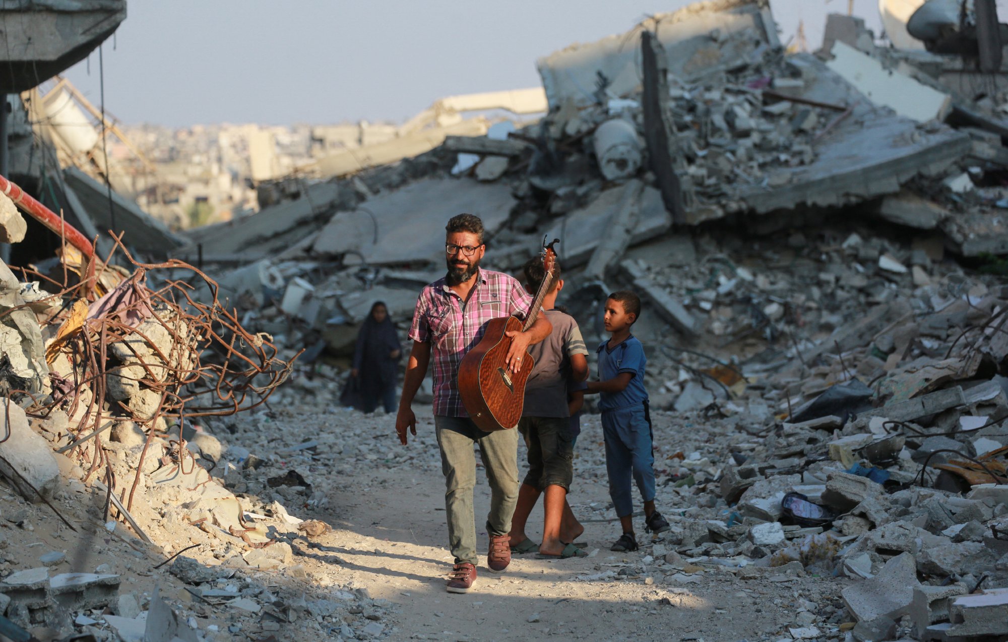L'immagine mostra un uomo che cammina tra le macerie di un'area distrutta, portando una chitarra. Sembra essere felice mentre si muove, accompagnato da un bambino. Sullo sfondo ci sono molti detriti e edifici in rovina, che suggeriscono una situazione di distruzione. Un'altra figura, probabilmente una donna, è visibile più lontano tra le macerie. L'atmosfera trasmette un contrasto tra la musica e la devastazione circostante.
