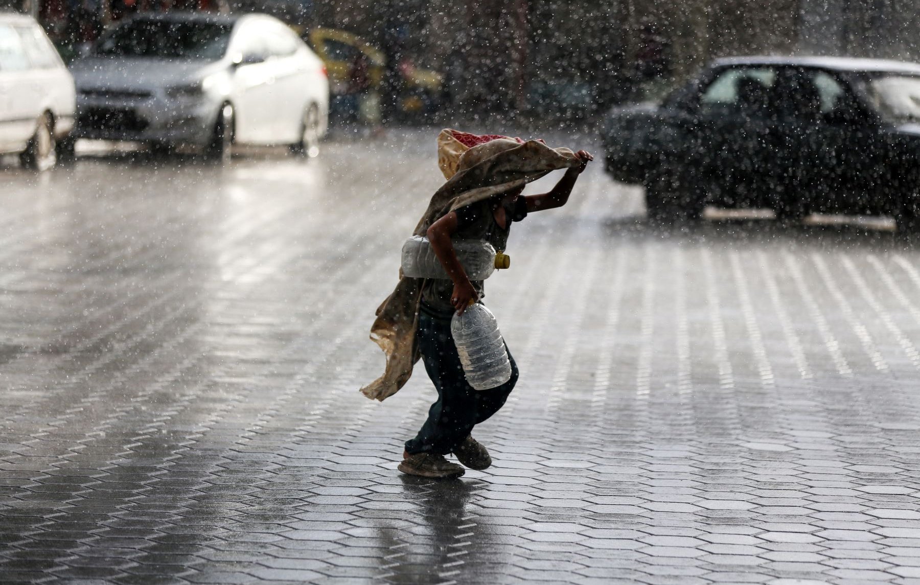 Nell'immagine si vede un bambino che corre sotto la pioggia intensa. Il ragazzo tiene un grande contenitore in una mano e si protegge dalla pioggia con un pezzo di stoffa. Sullo sfondo, ci sono diverse auto e una strada bagnata, mentre le gocce d'acqua creano un'atmosfera drammatica e vivace. La scena trasmette una sensazione di urgenza e resilienza, evidenziando il contrasto tra il bambino e le avversità del tempo.