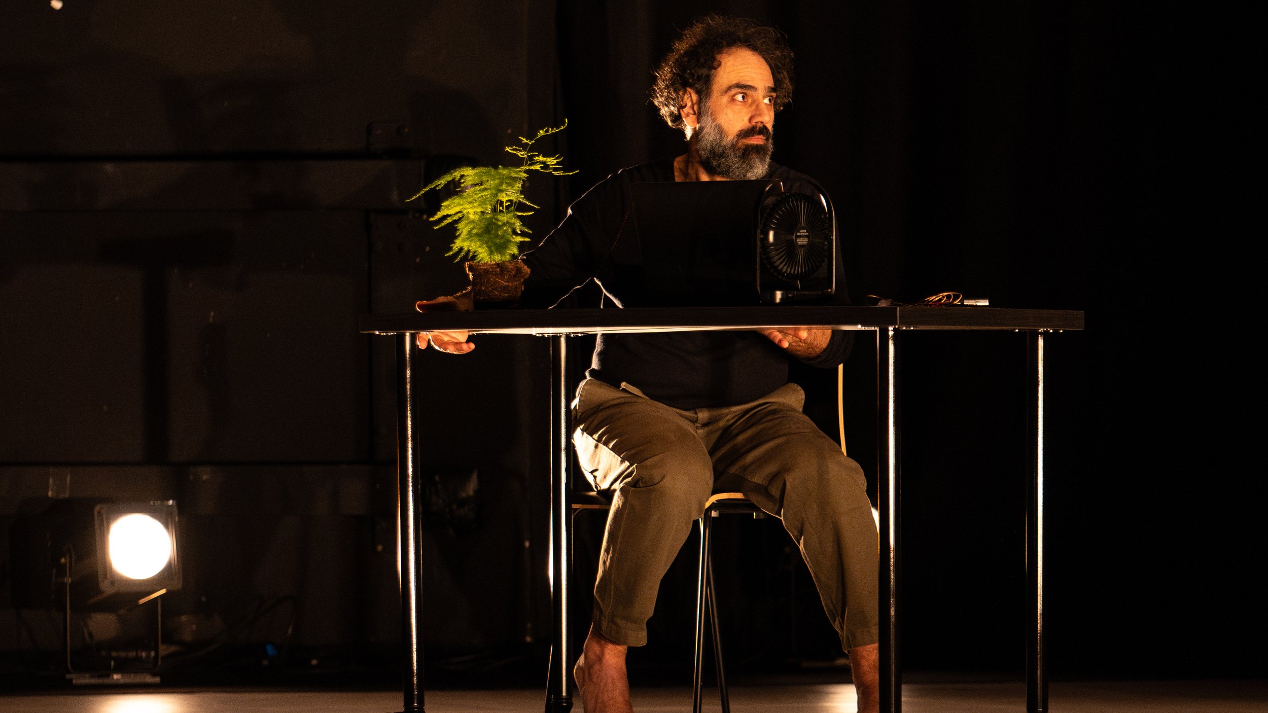 L'immagine mostra un uomo seduto a un tavolo in un ambiente di scena illuminato. Indossa una maglietta nera e pantaloni chiari, e sembra concentrato su un oggetto davanti a lui. Accanto a lui c'è una piccola pianta in vaso, mentre una ventola è posizionata sulla sua sinistra. L'illuminazione crea un'atmosfera drammatica, evidenziando sia l'uomo che gli oggetti sul tavolo. L'ambiente è scuro, con alcune luci che si riflettono sul pavimento.