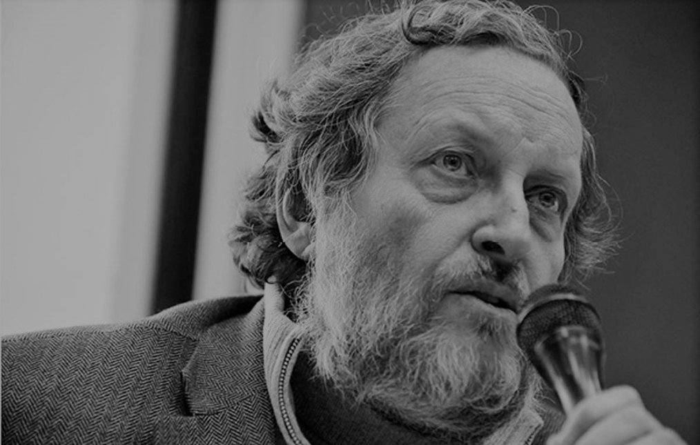 L'immagine mostra un uomo con un aspetto intellettuale. Ha una barba e capelli ricci, e sembra in fase di parlare al microfono. Indossa una giacca grigia e un maglione, trasmettendo un'aria di serietà e riflessione. Lo sfondo è sfocato, mettendo in evidenza il suo volto e la sua espressione concentrata.