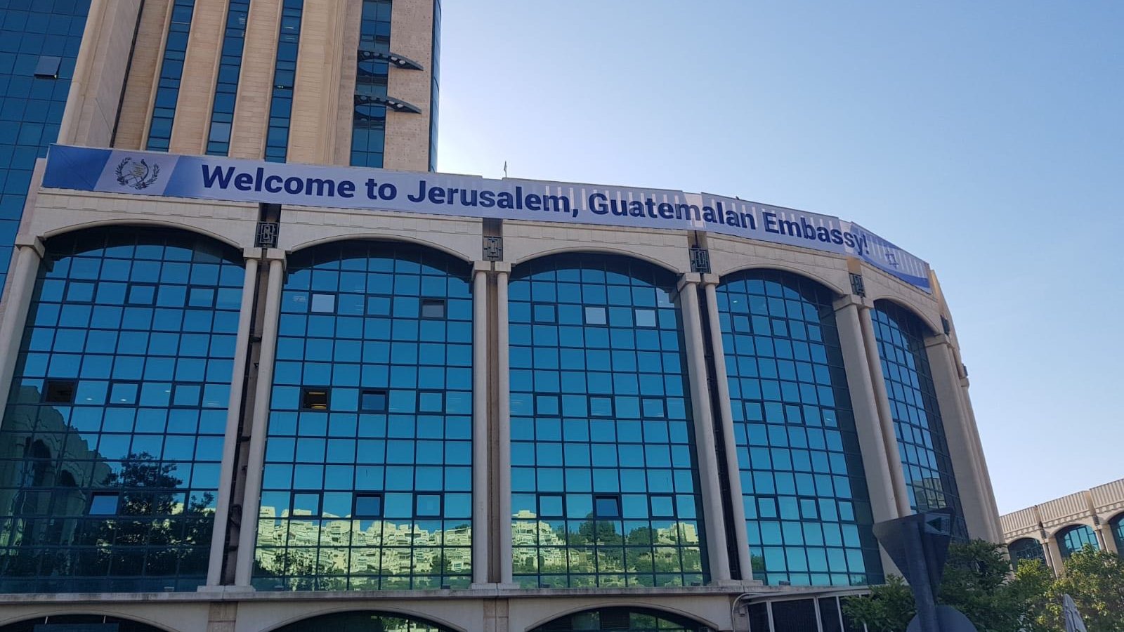 L'immagine mostra un edificio con una grande facciata di vetro riflettente. Sulla parte superiore dell'edificio è affisso uno striscione che dice "Welcome to Jerusalem, Guatemalan Embassy". Il contesto suggerisce che si tratta della sede dell'ambasciata del Guatemala a Gerusalemme. Si può notare il cielo blu sullo sfondo e alcune piante o alberi nella parte inferiore dell'immagine.