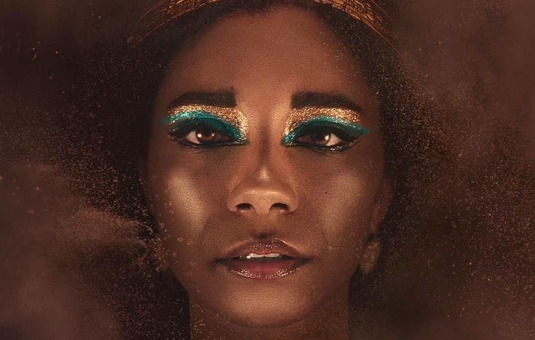 L'immagine presenta un volto femminile con un trucco molto elaborato, caratterizzato da ombretti blu e oro che mettono in risalto gli occhi. La persona indossa una corona decorata con serpenti dorati, che conferisce un aspetto regale e misterioso. Lo sfondo è sfocato e sembra essere avvolto da una polvere dorata, creando un'atmosfera affascinante e drammatica. La luce è calda e accentua la bellezza del viso, rendendo l'immagine suggestiva e intrigante.
