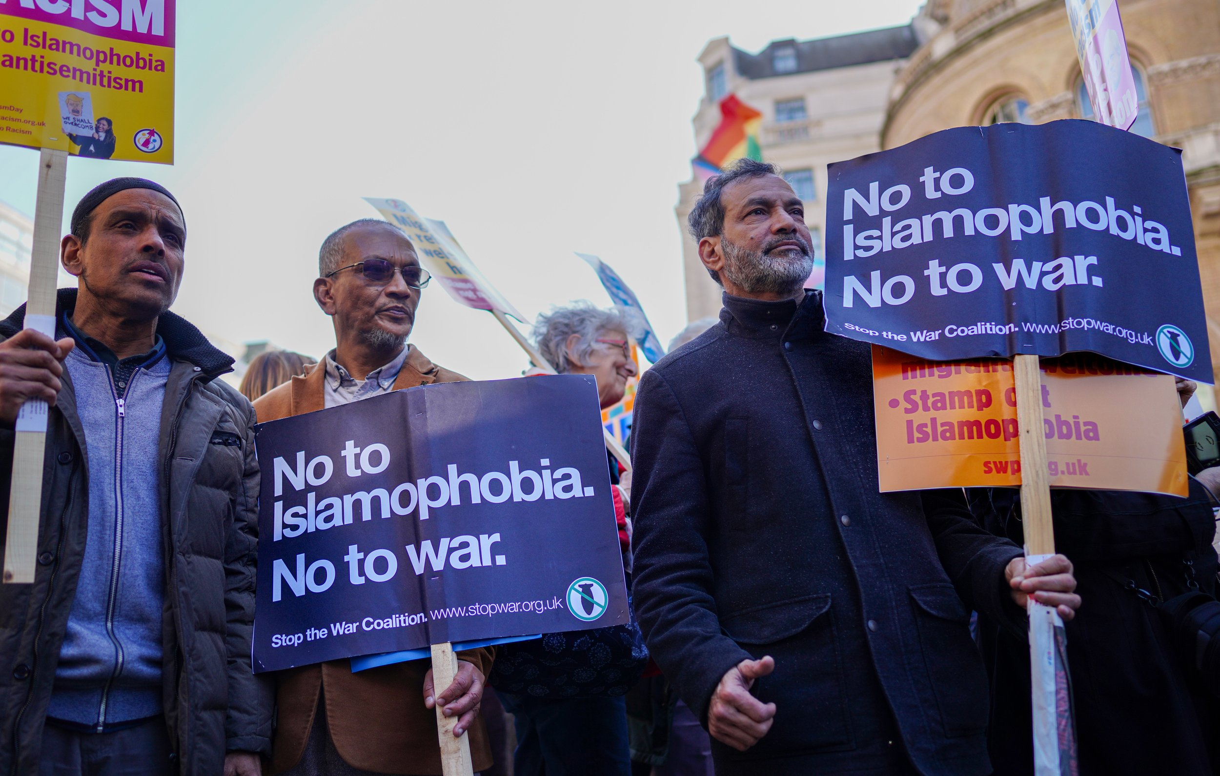 L'immagine mostra un gruppo di persone che partecipano a una manifestazione. Alcuni di loro stanno tenendo cartelli che riportano messaggi come "No to Islamophobia. No to war." e "Stop the War Coalition." L'atmosfera è di protesta pacifica, con i manifestanti che esprimono i loro punti di vista contro l'islamofobia e le guerre. In lontananza, si possono notare delle bandiere, che aggiungono un senso di diversità e unità all'evento.
