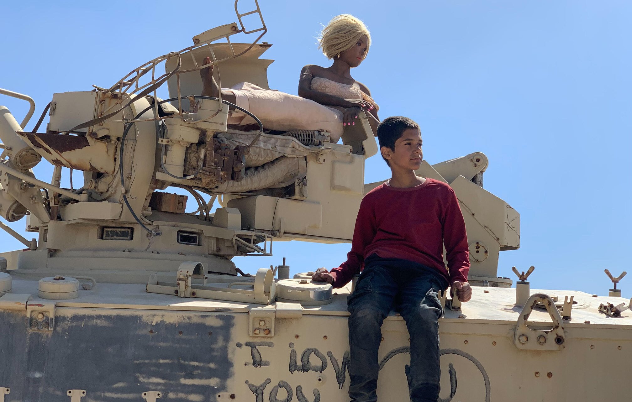 L'immagine mostra un giovane ragazzo seduto su un veicolo militare, apparentemente un carro armato. Accanto a lui c'è una figura che sembra essere una bambola o un manichino con capelli biondi. Sul carro armato è visibile una scritta che dice "I love you". Il cielo è limpido e blu, creando un contrasto con l'ambiente circostante. Il ragazzo indossa un maglione rosso e jeans, mentre la bambola è posizionata in modo da sembrare tranquilla e serena.