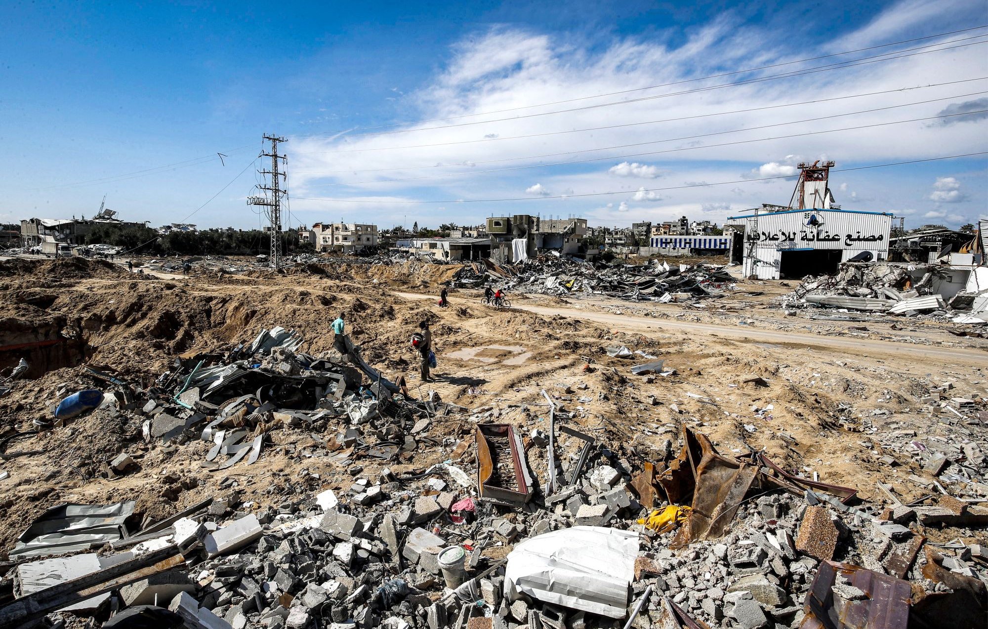 L'immagine mostra un paesaggio devastato, caratterizzato da macerie e detriti. Il terreno sembra essere stato scavato e distrutto, con edifici in rovina visibili sullo sfondo. I resti di strutture crollate sono sparsi ovunque, mentre alcune persone si muovono tra le macerie. Il cielo è parzialmente nuvoloso, e ci sono segnali di infrastrutture elettriche nei dintorni. L'atmosfera trasmette un senso di desolazione e distruzione.