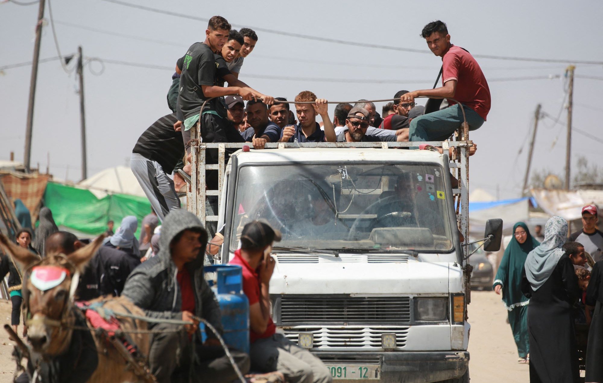 L'immagine mostra un gruppo di persone che si trovano su un camion, alcuni di loro sono seduti sul retro del veicolo, mentre altri sono in piedi. Sul lato destro ci sono persone su un carro trainato da un animale, che sembra essere un cavallo. In fondo si intravedono tende e strutture, suggerendo un contesto urbano o di accampamento. L'atmosfera sembra vivace, con diverse persone che interagiscono tra loro.