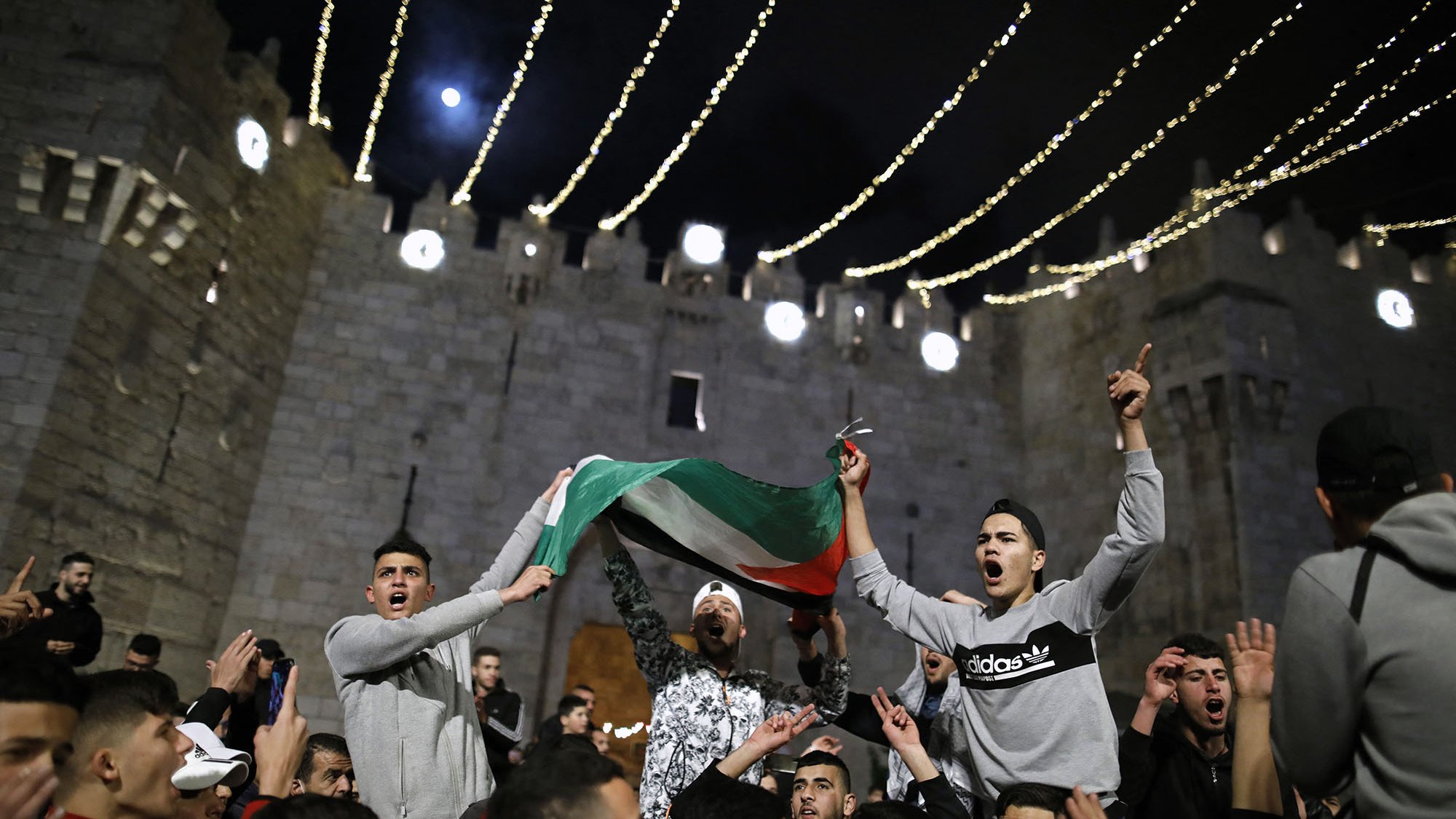 L'immagine mostra un gruppo di persone riunite in un contesto festivo, probabilmente in un evento pubblico. I partecipanti stanno sventolando una bandiera, che sembra essere quella palestinese, e sono visibilmente entusiasti. Sullo sfondo, si può notare una struttura medievale, che suggerisce un'atmosfera di celebrazione. Le luci appese creano un'atmosfera suggestiva e notturna, mentre i volti delle persone esprimono gioia e partecipazione.