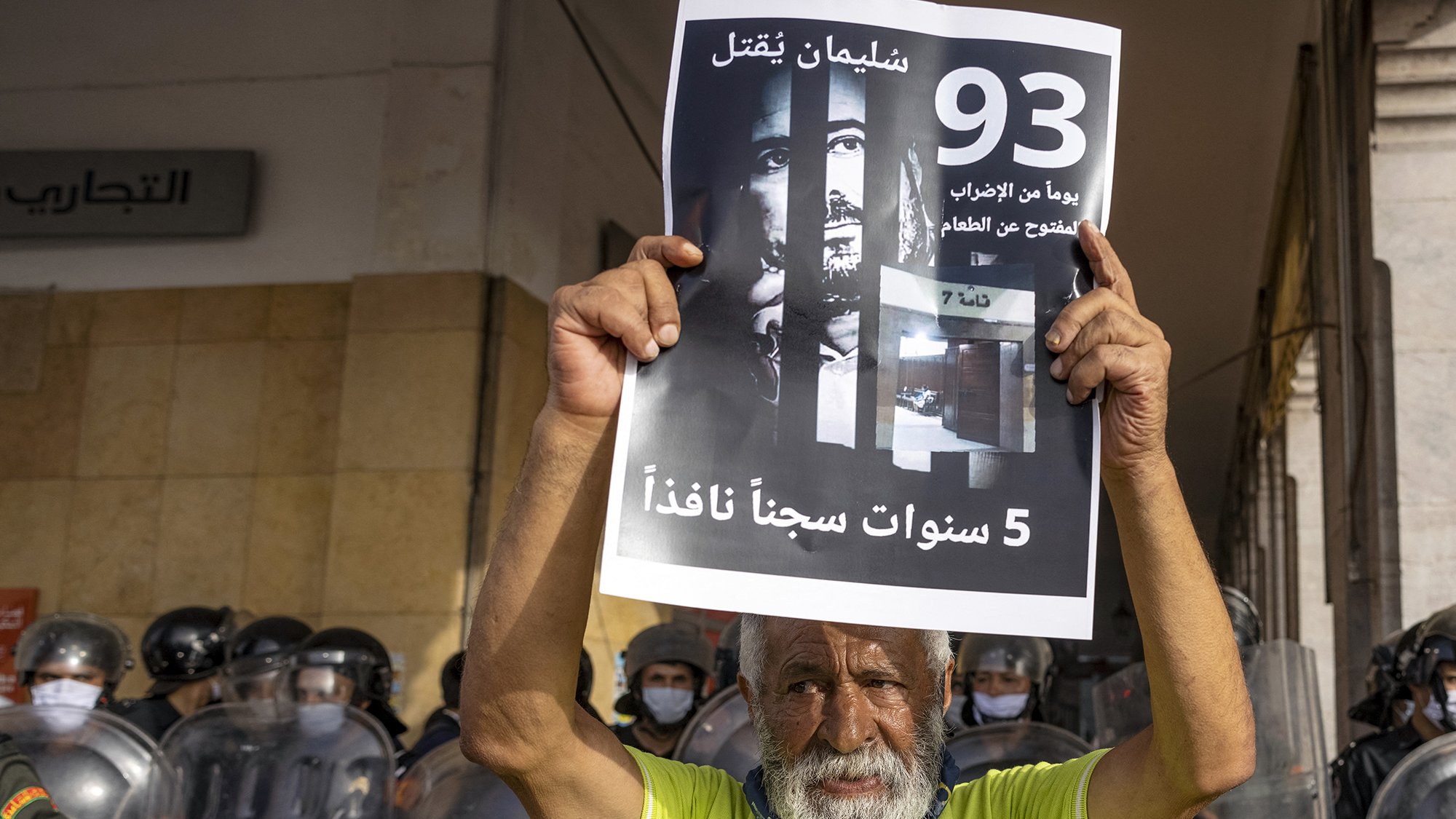 L'immagine mostra un uomo anziano che tiene in alto un cartello. Sul cartello si possono vedere scritte in arabo e un'immagine di volti, che suggerisce un tema di protesta o di richiesta di giustizia. Sullo sfondo ci sono agenti di polizia in uniforme antisommossa, suggerendo che l'evento si svolge in un contesto di tensione. L'anziano sembra determinato mentre partecipa a questa manifestazione.