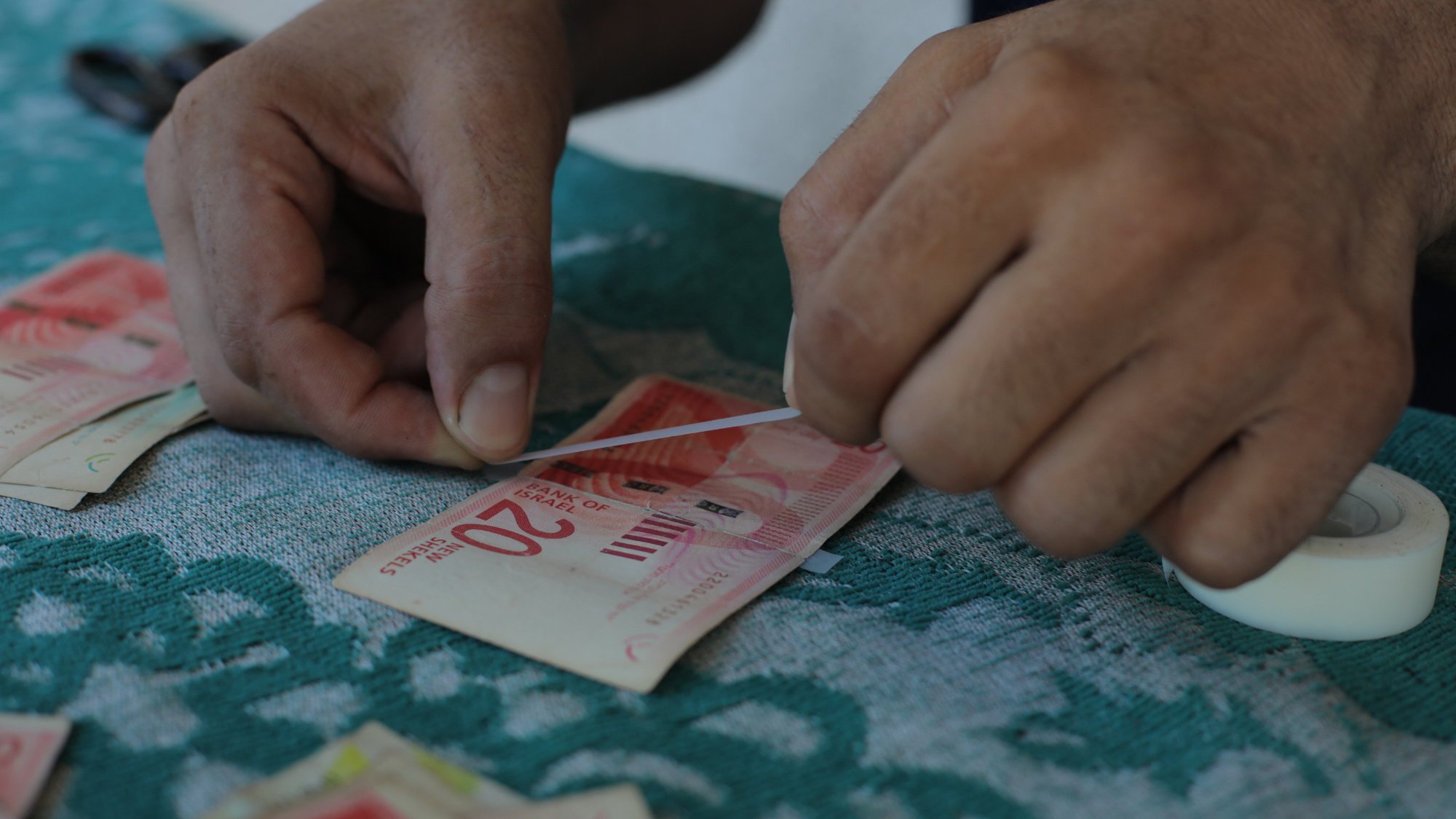 L'immagine mostra delle mani che stanno piegando e preparando delle banconote, probabilmente di grosso taglio. Le banconote sono adagiate su un tavolo con una superficie di tessuto verde, e accanto si vede un rotolo di nastro adesivo. I movimenti delle mani suggeriscono che si sta cercando di raggruppare o sistemare il denaro in modo ordinato.