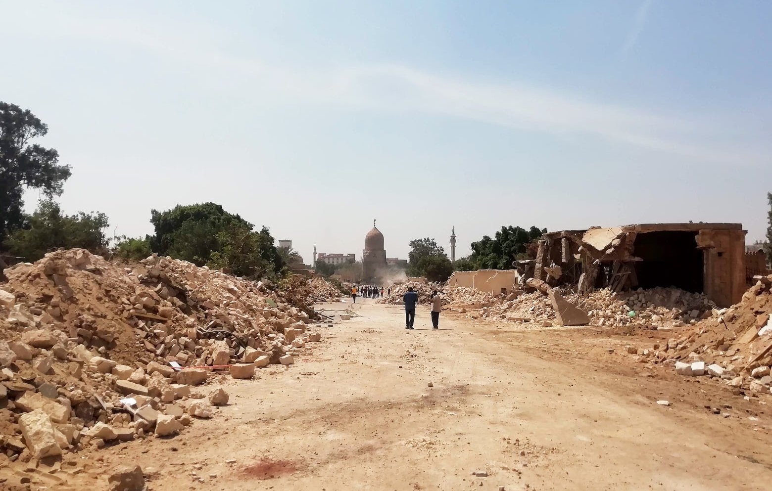 L'immagine mostra una strada sterrata, circondata da macerie e detriti. Sullo sfondo si intravede un edificio con una cupola, probabilmente una moschea o un monumento, avvolto da polvere. La scena trasmette un senso di devastazione, con alberi e strutture danneggiate ai lati della strada. Alcune persone sono visibili in lontananza, mentre il cielo appare chiaro e luminoso.