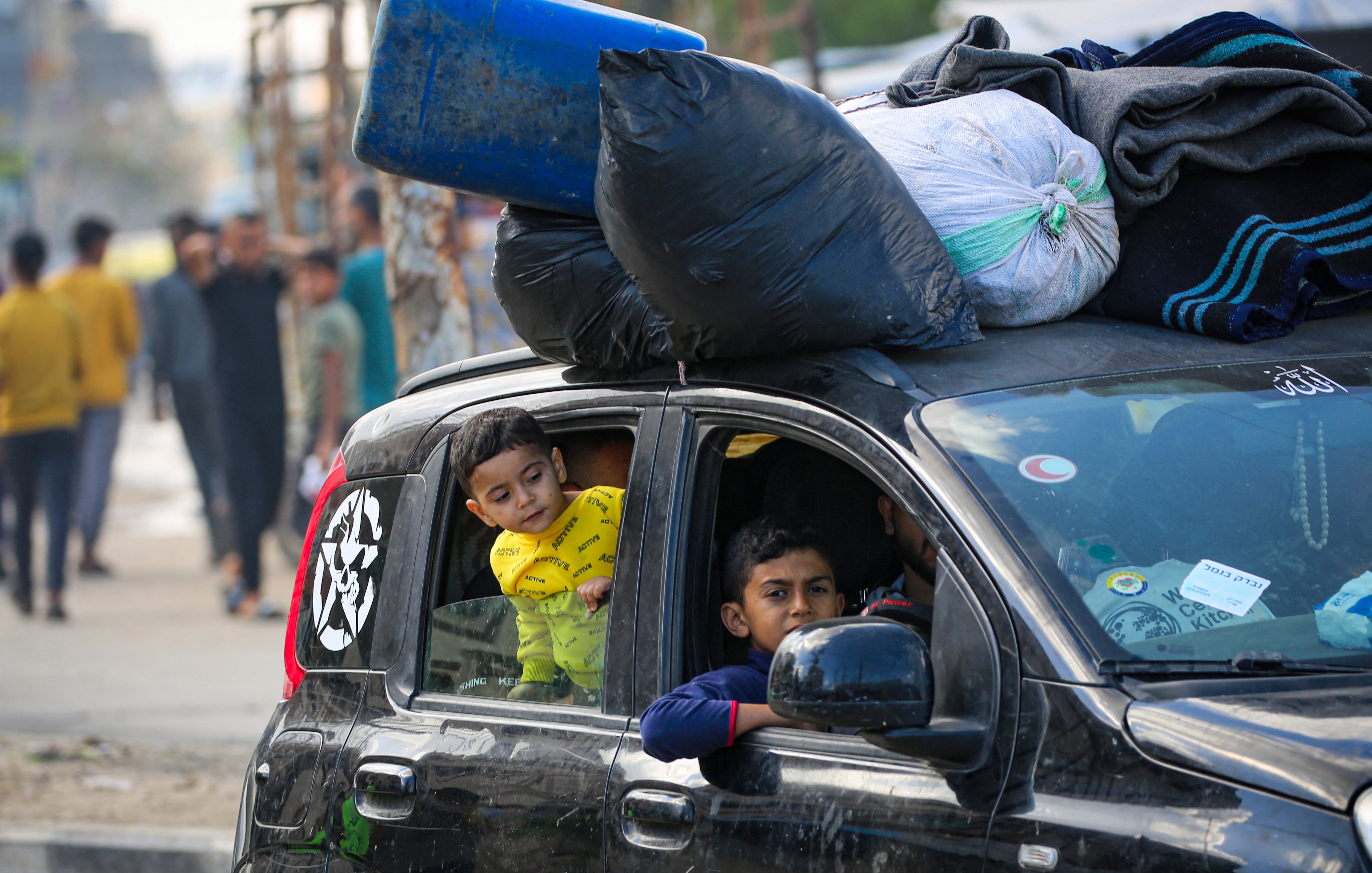 L'immagine mostra un'auto con un carico sopra di sacchi neri e altri oggetti. All'interno della vettura, si vedono due bambini: uno che guarda fuori dal finestrino con espressione curiosa e l'altro che si affaccia dall'apertura. Sullo sfondo si possono notare altre persone e un ambiente urbano, che sembra un'area affollata. La scena riflette una situazione di vita quotidiana in un contesto urbano.