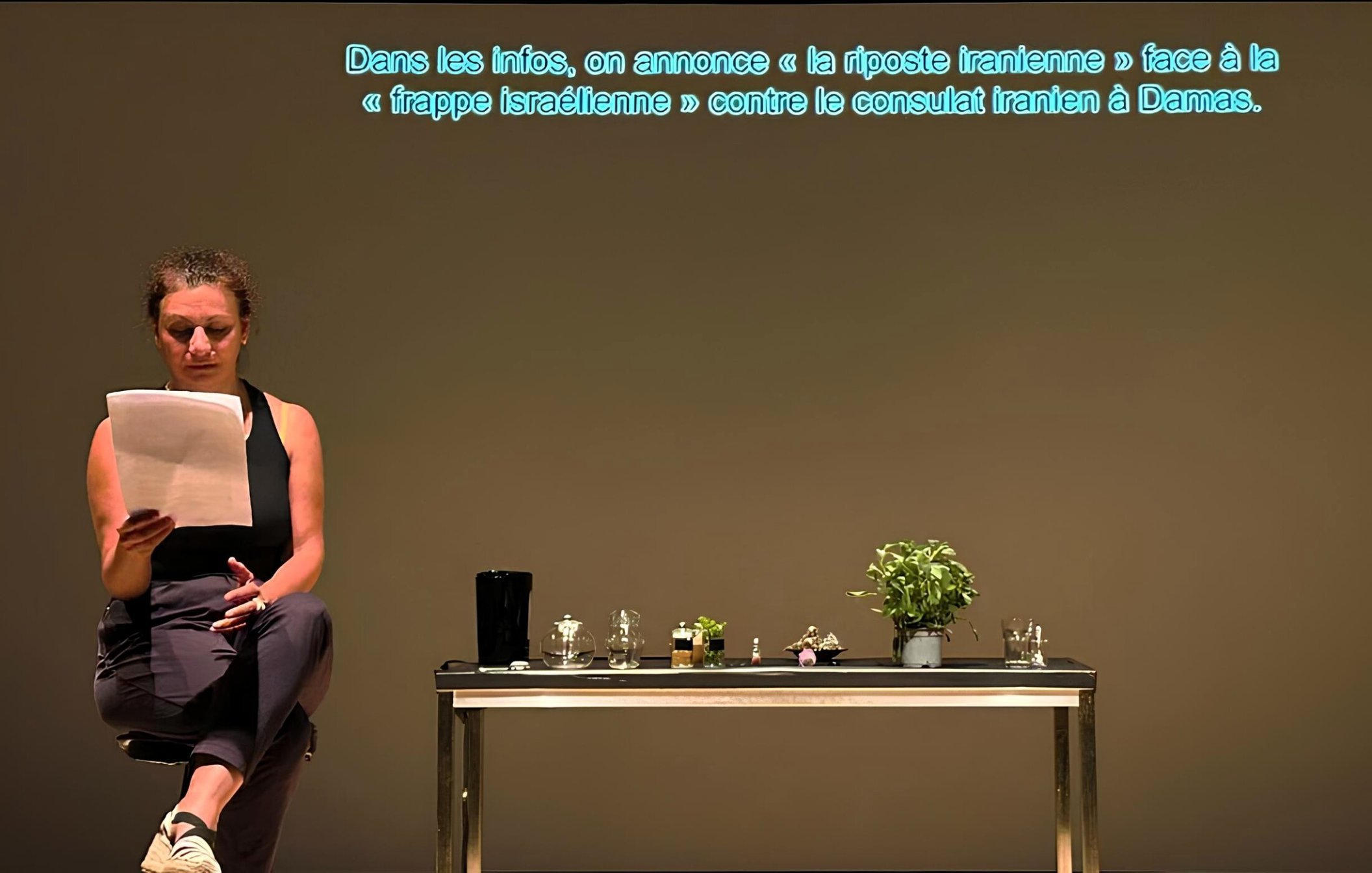 Una donna legge appunti davanti a un tavolo con piante e oggetti, su uno sfondo neutro.