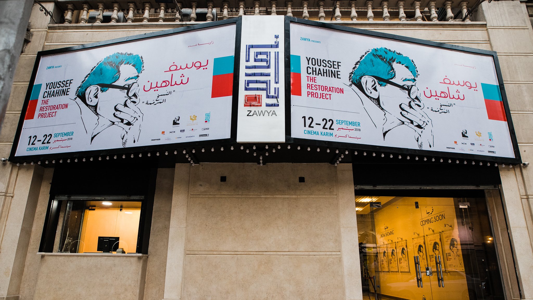 L'immagine mostra un edificio con una facciata decorata da grandi manifesti. I manifesti annunciano un progetto artistico intitolato "The Restoration Project" di Youssef Chahine, con date di evento indicate dal 12 al 22 settembre. Si può notare un design grafico accattivante, con illustrazioni e testo in arabo e in inglese. La zona sembra essere un luogo di cultura o un centro espositivo, con elementi architettonici tipici di un contesto urbano.