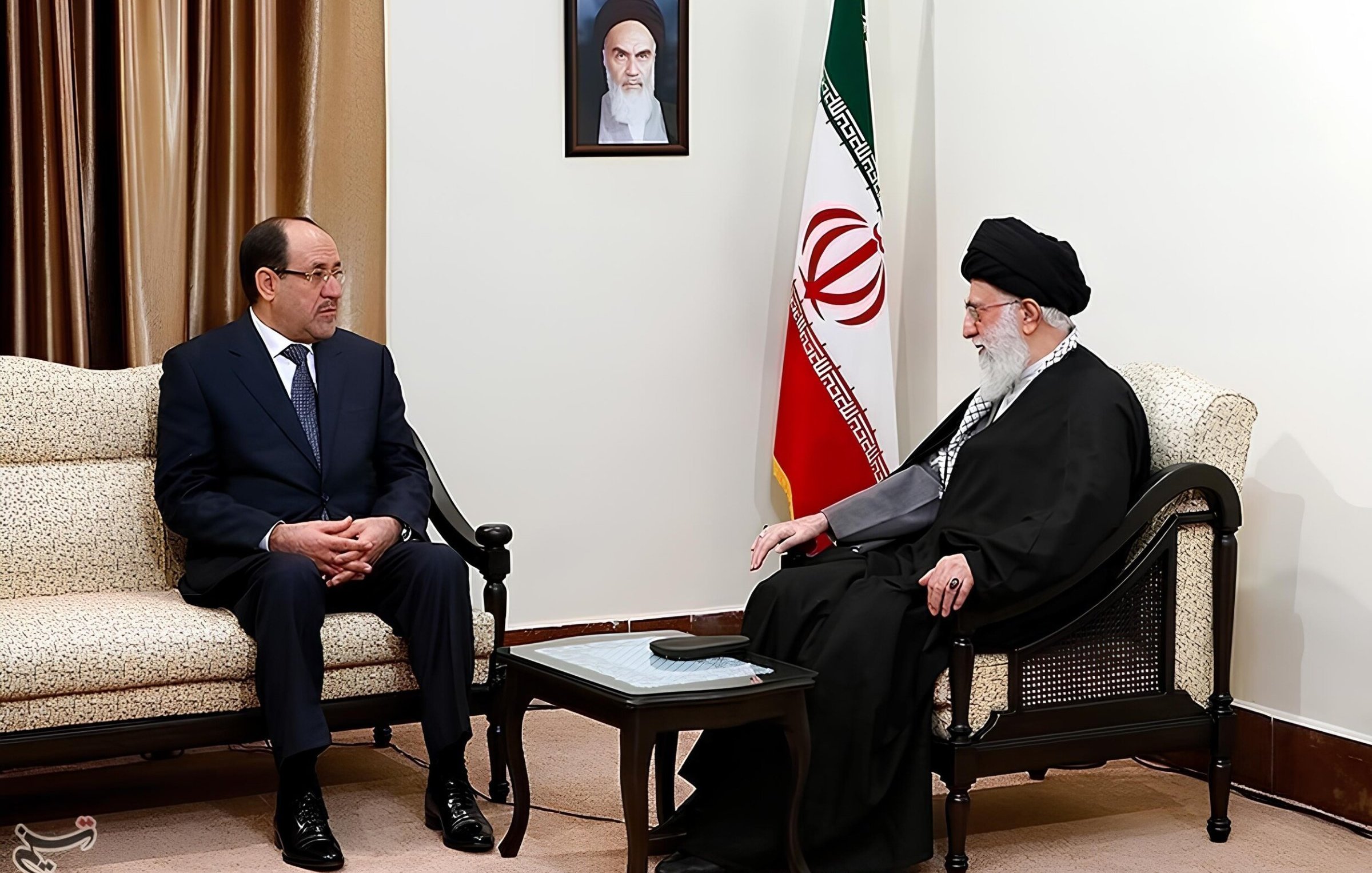 Due uomini seduti in una sala con bandiera iraniana, discutono seriamente.