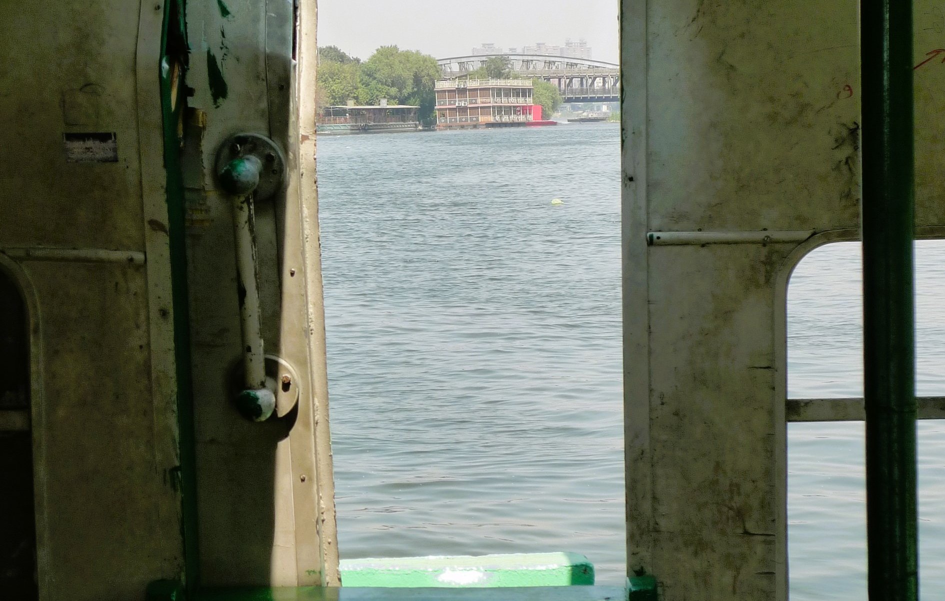 L'immagine mostra l'interno di un'imbarcazione, con una porta aperta che si affaccia su un corpo d'acqua. La vista rivela un paesaggio sereno, con un riflesso dell'acqua e vegetazione visibile in lontananza. La struttura della barca appare semplice, con pareti in metallo e sedili visibili all'interno. La luce naturale entra attraverso l'apertura, creando un'atmosfera tranquilla.