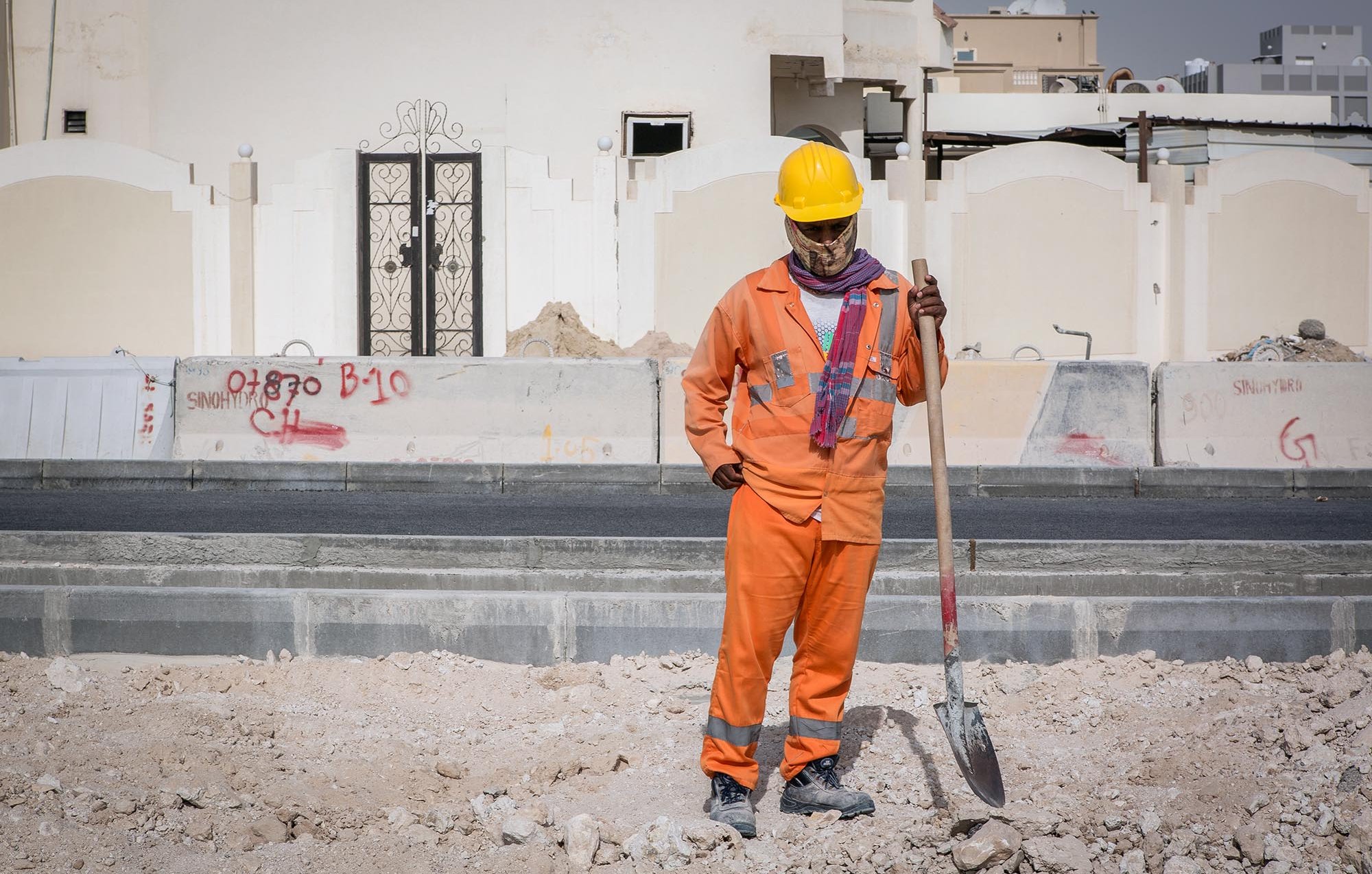 Nell'immagine si vede un lavoratore in un cantiere edile. Indossa una tuta arancione e un casco giallo per la sicurezza. Ha in mano una pala e sembra essere in pausa, osservando il terreno di fronte a lui. Sullo sfondo si possono notare alcune strutture e graffiti su un muro. L'ambiente appare polveroso e in fase di costruzione.