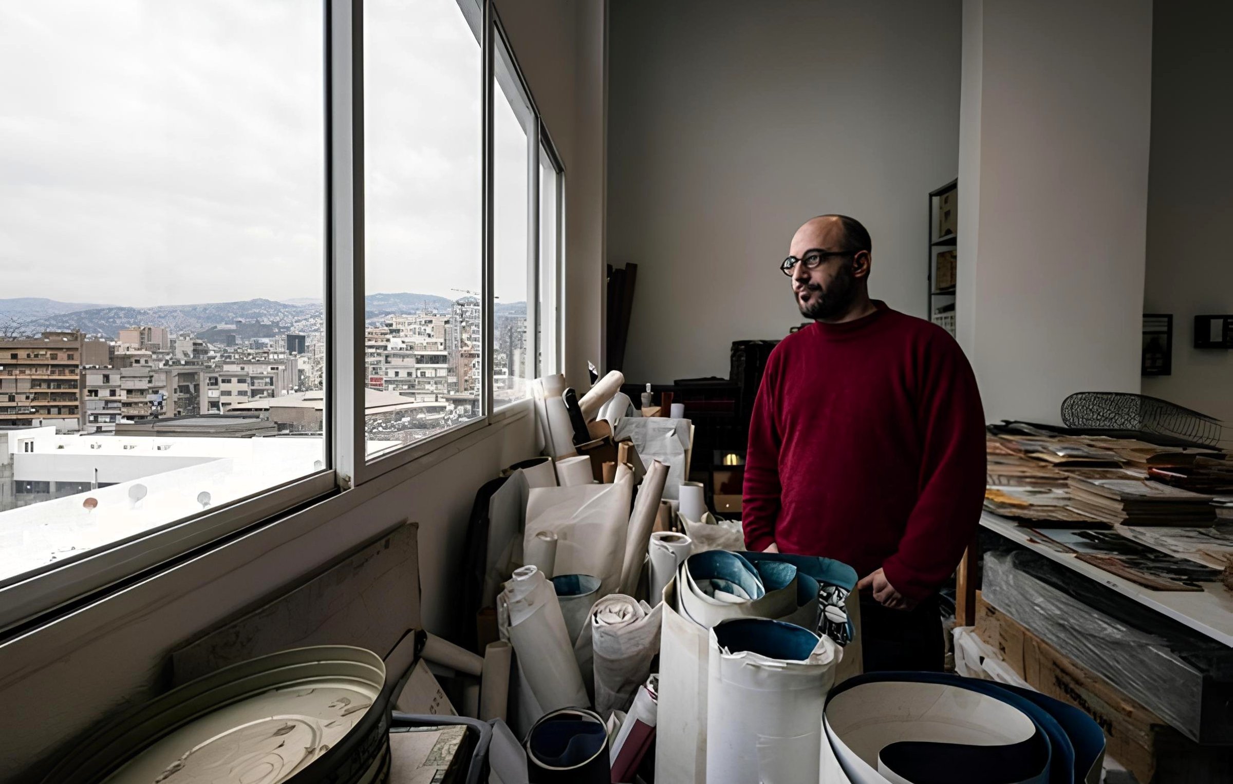 Uomo in maglione rosso guarda fuori da una finestra, circondato da rotoli di carta e materiali.