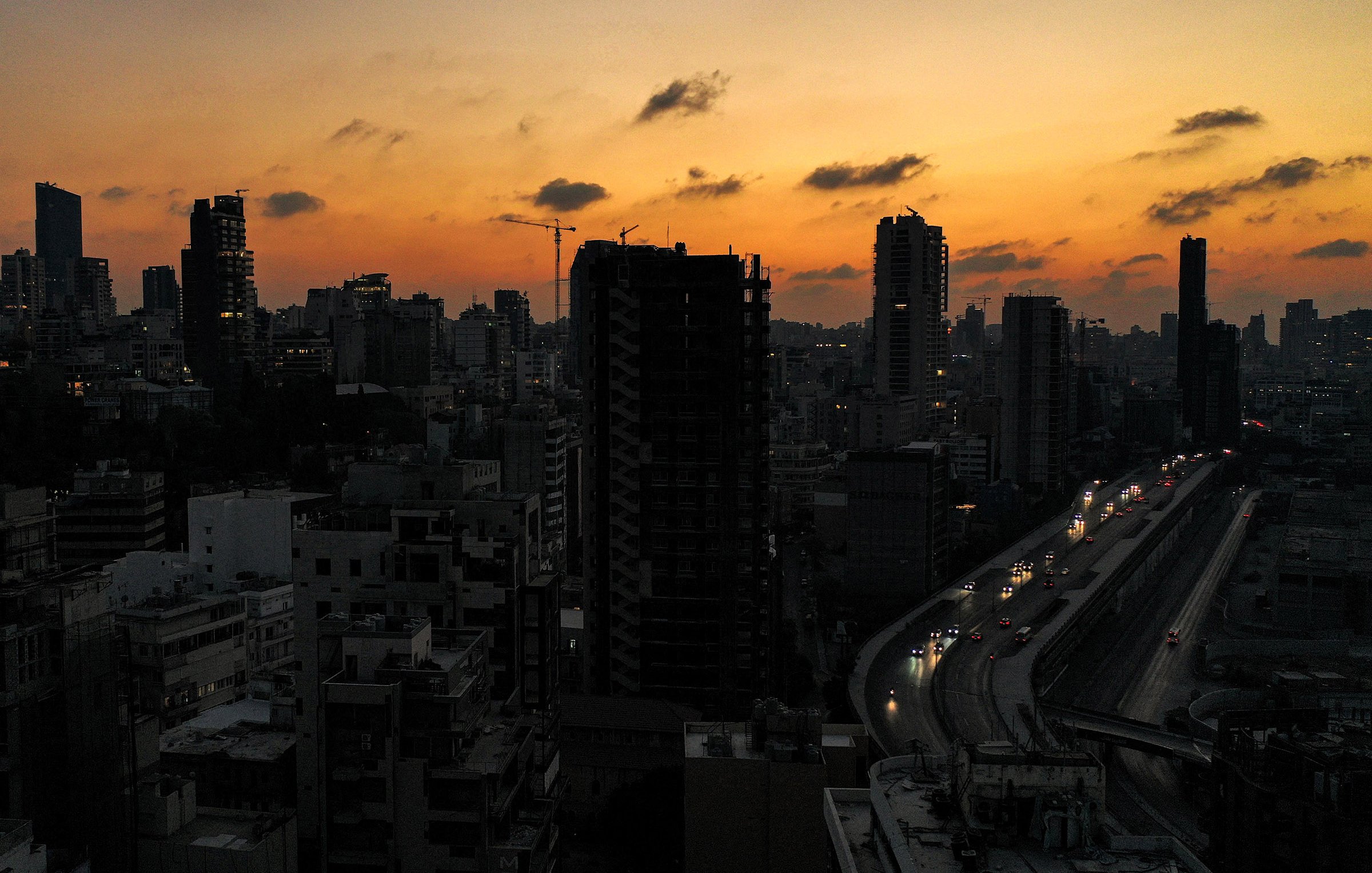 L'immagine ritrae un paesaggio urbano al tramonto, con il cielo che mostra tonalità calde di arancione e giallo. Silhouette di grattacieli si stagliano contro il cielo, creando un contrasto tra il buio degli edifici e la luce del tramonto. In basso, si vede una strada con auto che circolano, illuminata dalle luci dei veicoli. L'atmosfera è serena e suggestiva, tipica di un momento di transizione tra giorno e notte.