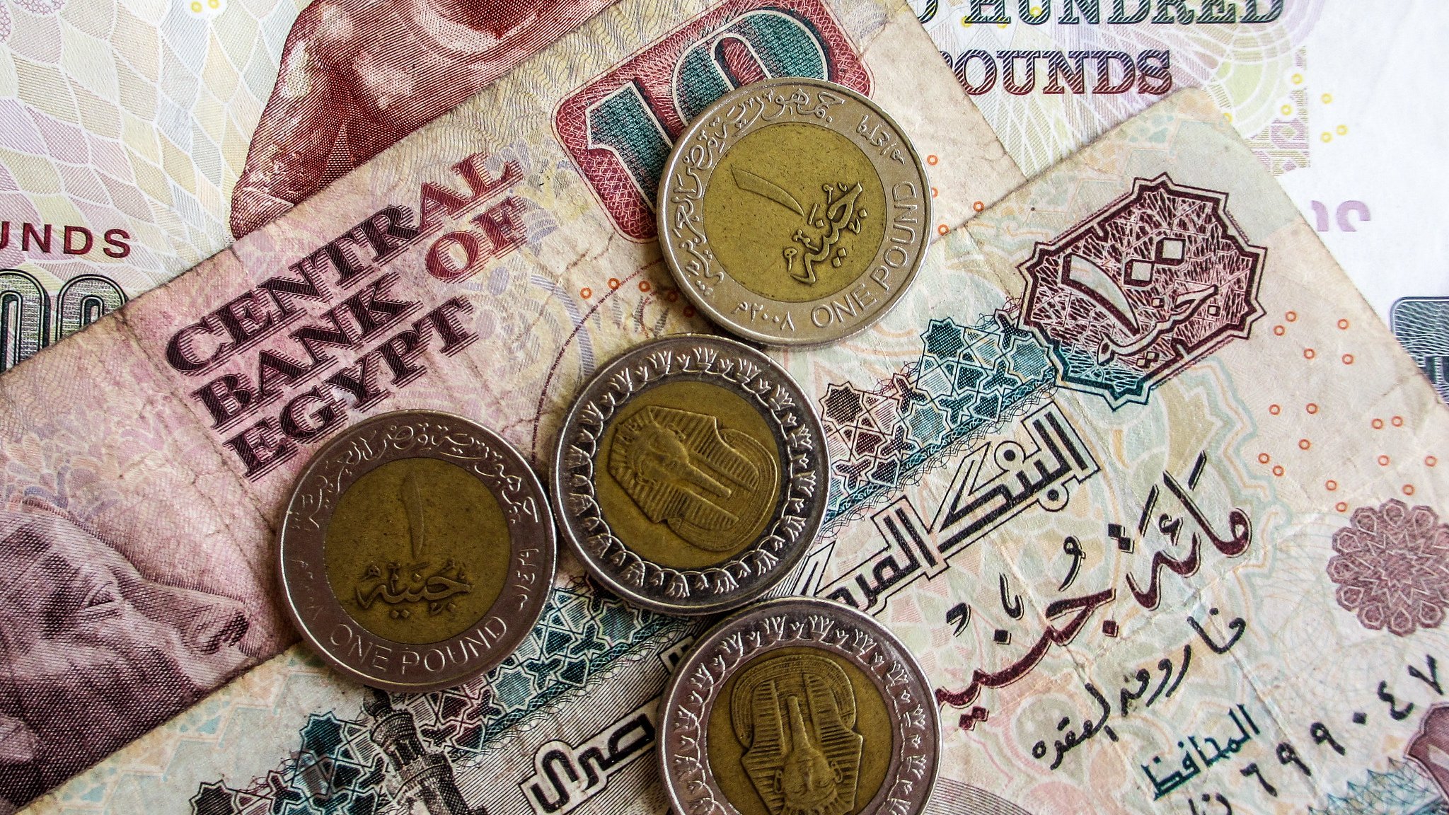 L'immagine mostra diverse banconote e monete egiziane. In primo piano ci sono quattro monete, due delle quali sono di un pound egiziano. Sullo sfondo, ci sono banconote di diverse denominazioni, evidentemente con scritte arabe, tipiche della valuta egiziana. Una banconota sembra avere il valore di cento pounds e presenta un motivo decorativo. L'atmosfera è quella di un contesto economico, evidenziando elementi tipici della valuta dell'Egitto.