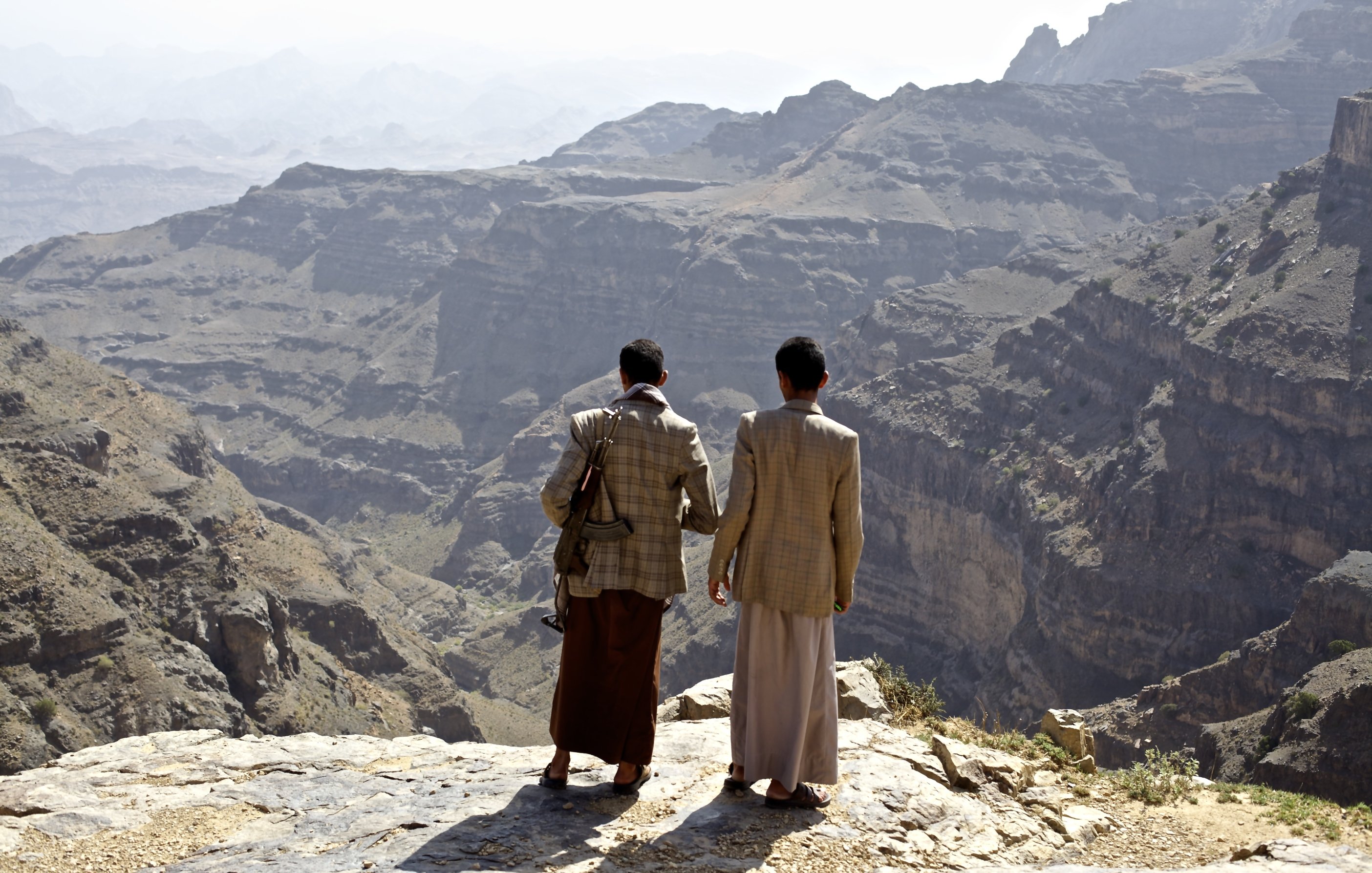 L'immagine mostra due uomini che si trovano su un bordo roccioso, ammirando un vasto paesaggio montano. Le loro spalle sono visibili mentre guardano verso il fondo di una valle profonda e frastagliata. Le montagne circostanti sono di diverse tonalità di grigio e marrone, con una vegetazione scarsa visibile in alcune zone. L'atmosfera appare serena e suggestiva, con un cielo chiaro sullo sfondo.