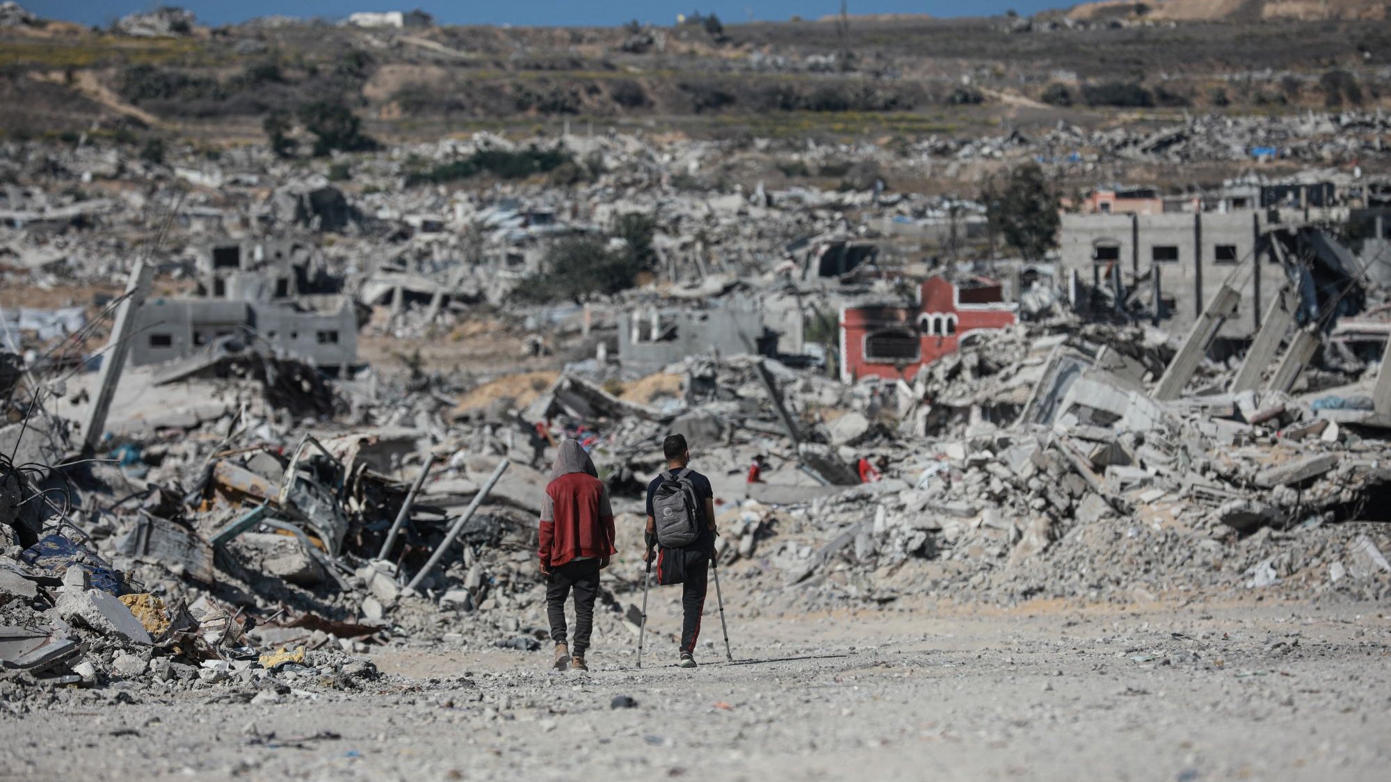 Due persone camminano tra le macerie di edifici distrutti in un paesaggio desolato.