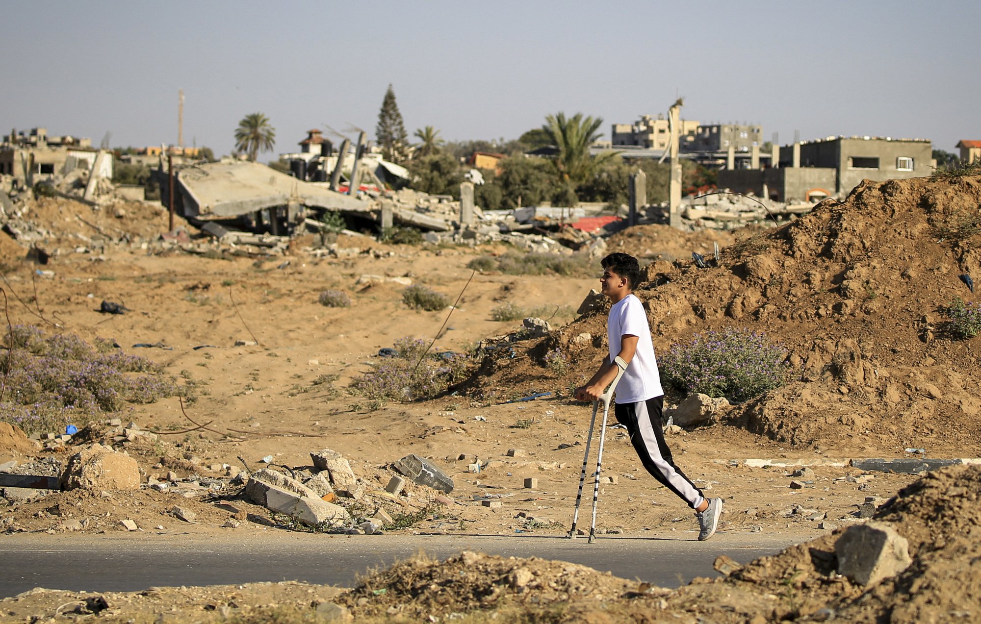 L'immagine mostra un giovane che cammina con le stampelle in un'area distrutta, circondata da macerie e detriti. Sullo sfondo si possono vedere edifici in rovina e un paesaggio desolato, con sabbia e vegetazione sparsa. La scena trasmette un senso di difficoltà e resilienza in un contesto di devastazione.