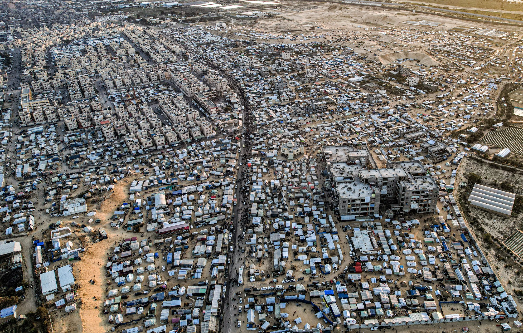 L'immagine mostra un'ampia area urbana vista dall'alto, caratterizzata da una fitta aggregazione di edifici e strutture. Si nota una forte densità abitativa, con molte abitazioni di dimensioni varie disposte in modo irregolare. Le strade principali e i passaggi sono visibili tra le varie costruzioni. In alcune zone si possono notare anche tende o strutture provvisorie, suggerendo la presenza di comunità temporanee. Sullo sfondo, si intravedono aree di terreni aperti e sviluppo urbano in espansione. La scena complessiva trasmette una sensazione di vivacità e organizzazione spontanea, con una tipologia abitativa che riflette una realtà complessa.