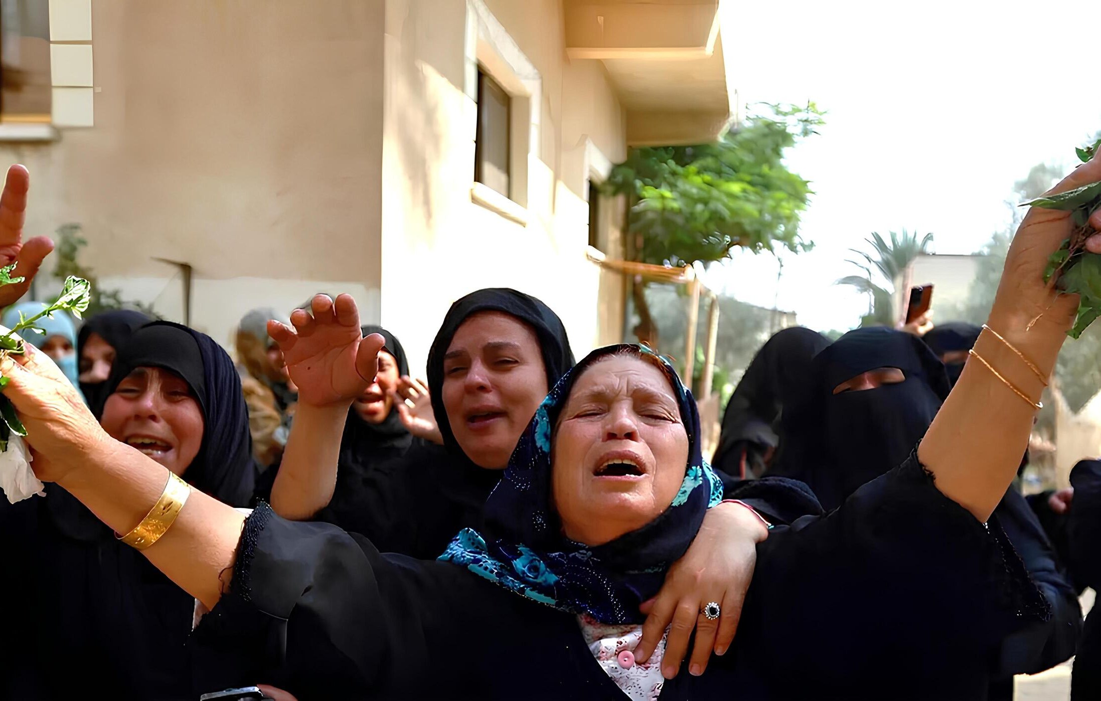 L'immagine mostra un gruppo di donne in un contesto emotivo intenso. Indossano abiti scuri e alcune di loro hanno il viso coperto. Alcune donne sembrano piangere e sollevano le braccia, mentre altre mostrano espressioni di sofferenza. In mano hanno foglie verdi, che possono rappresentare un simbolo di lutto o commemorazione. Sullo sfondo, si intravedono edifici e vegetazione. L'atmosfera è carica di emotività e partecipazione.