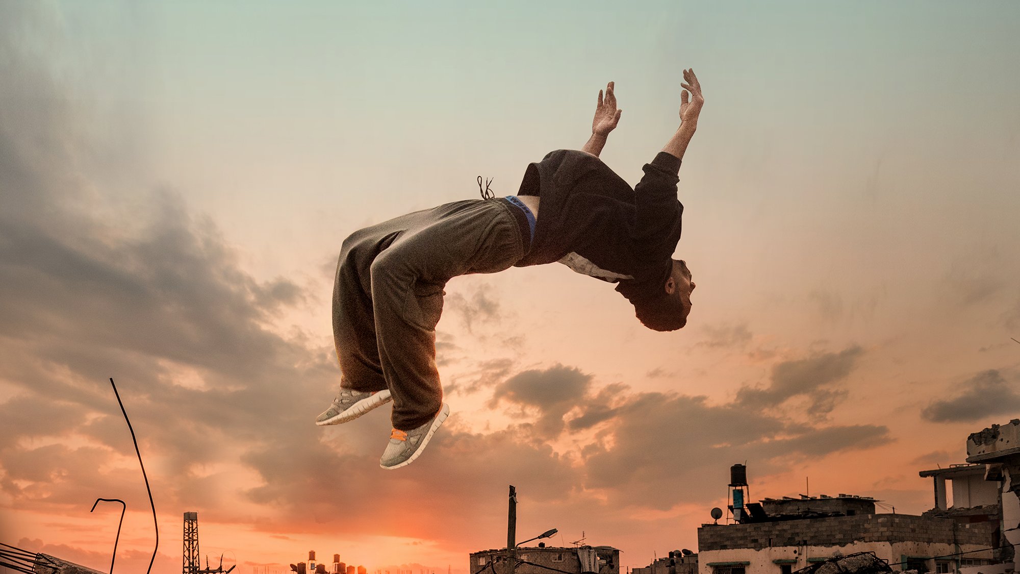L'immagine mostra un giovane che sembra fluttuare o saltare sopra un paesaggio urbano distrutto, con scarti di costruzione e rovine. Sullo sfondo, si possono vedere nuvole e un cielo al tramonto, che creano un'atmosfera drammatica. La silhouette del ragazzo è evidenziata dalla luce, evidenziando un senso di libertà e movimento in un contesto desolato.