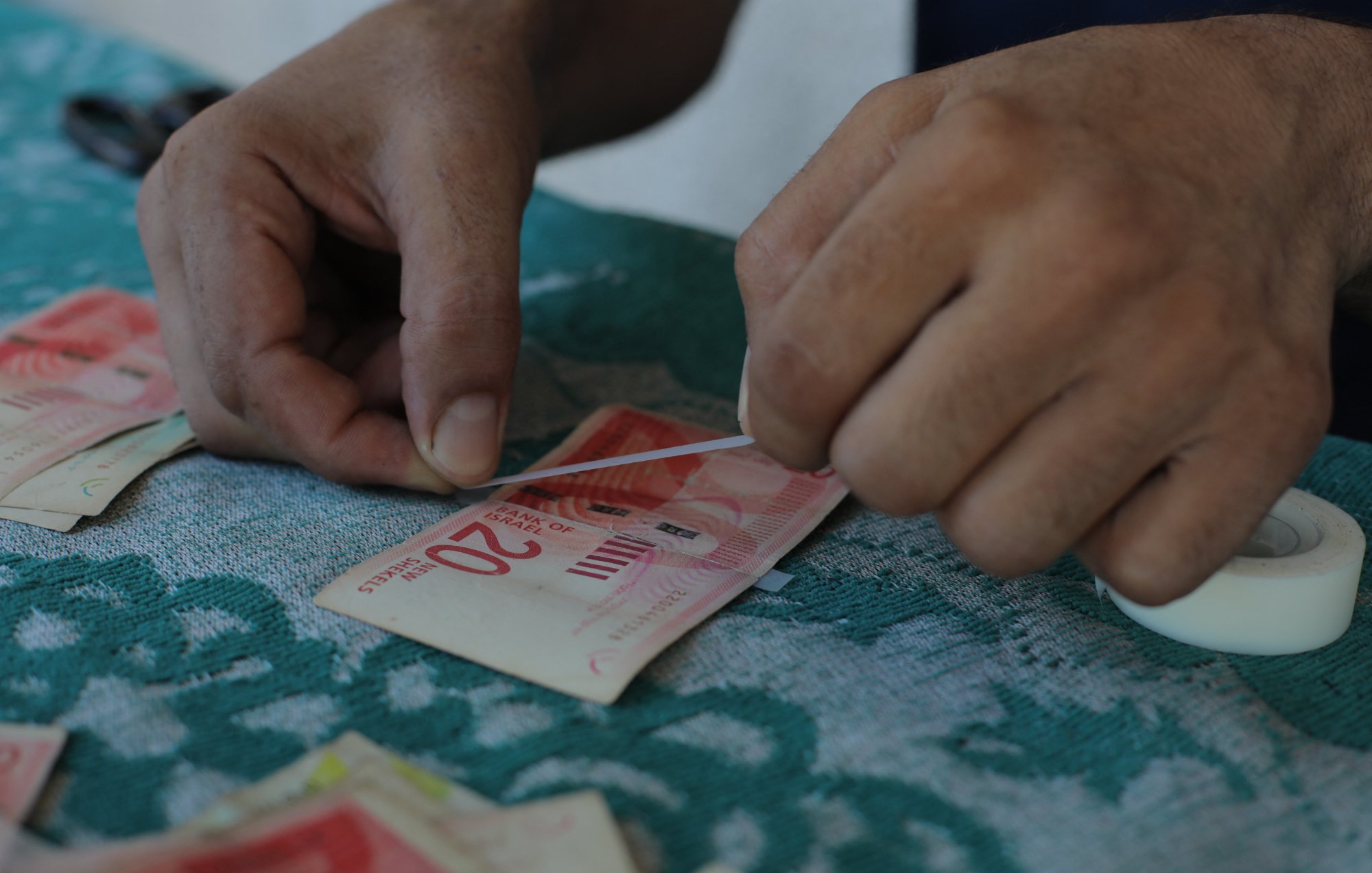 L'immagine mostra delle mani che stanno piegando e preparando delle banconote, probabilmente di grosso taglio. Le banconote sono adagiate su un tavolo con una superficie di tessuto verde, e accanto si vede un rotolo di nastro adesivo. I movimenti delle mani suggeriscono che si sta cercando di raggruppare o sistemare il denaro in modo ordinato.