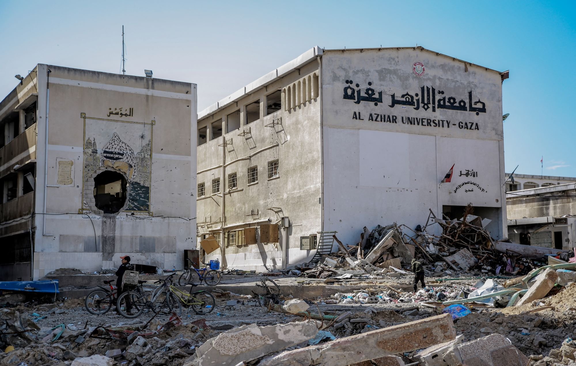 L'immagine mostra un edificio in rovina, probabilmente parte dell'Università Al Azhar di Gaza. La struttura presenta segni di distruzione, con macerie sparse e un'atmosfera di desolazione. A sinistra, si può notare un affresco o un murales deteriorato. Due persone si trovano nel campo visivo, una delle quali è in bicicletta. La scena riflette i danni significativi causati da conflitti e la situazione difficile in quella regione.