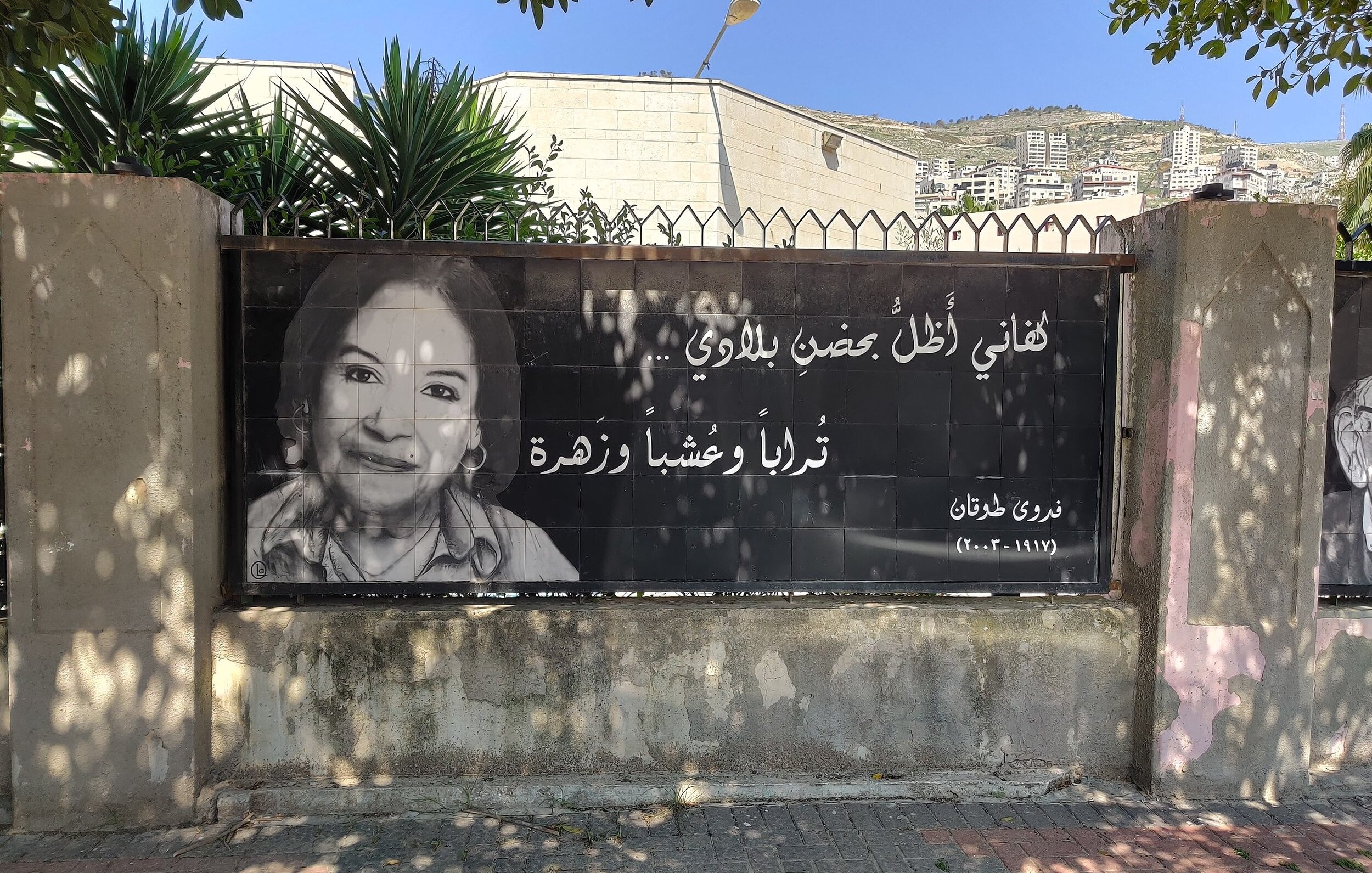 L'immagine mostra un grande striscione che riporta una fotografia di una donna sorridente, accompagnata da un testo in arabo. Il contesto sembra essere un'area urbana, con edifici sullo sfondo e piante verdi nelle vicinanze. L'atmosfera è accogliente e il messaggio sullo striscione appare positivo e invitante.